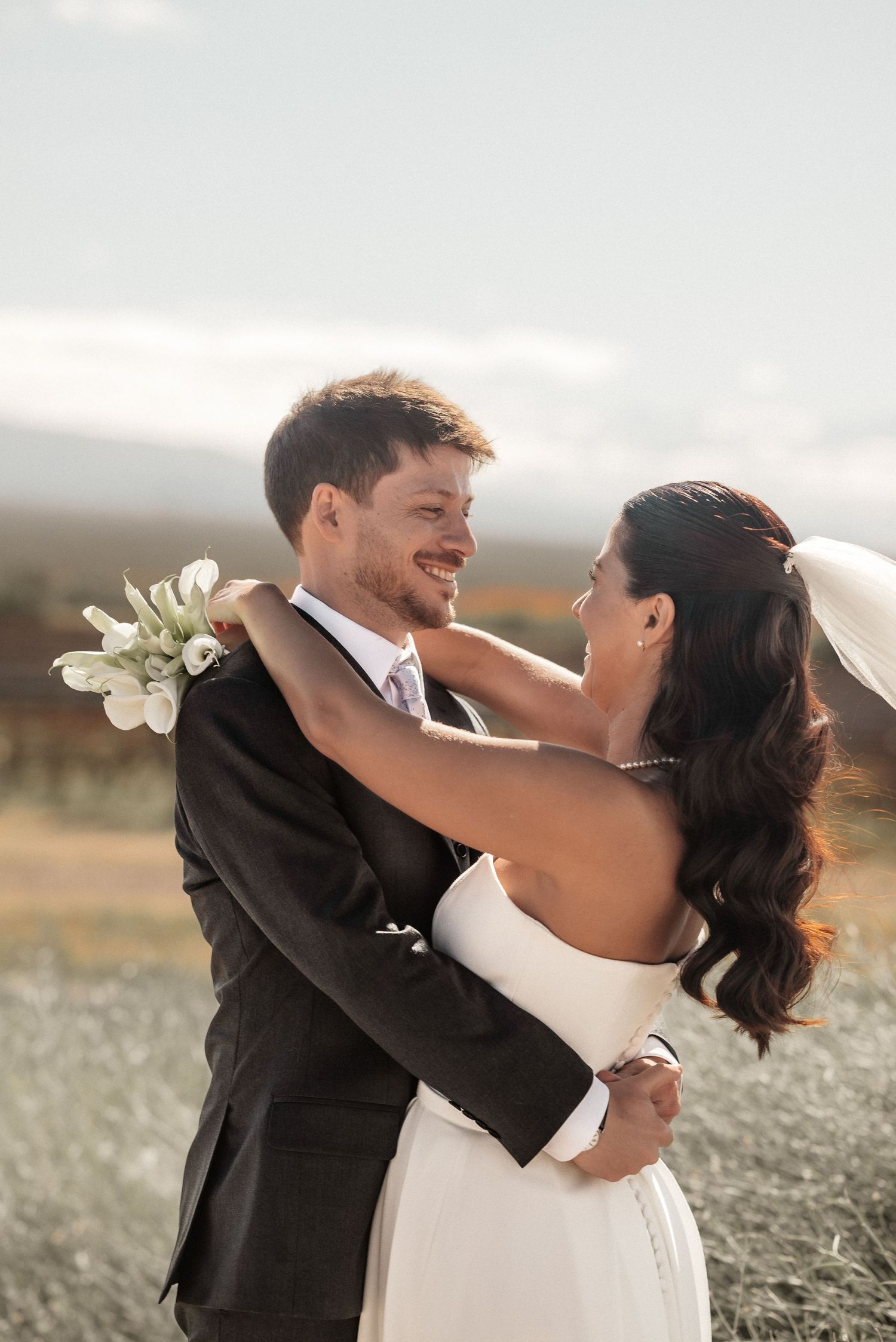 A smiling groom in a dark suit embraces his bride in a white dress and veil outdoors at their wedding.