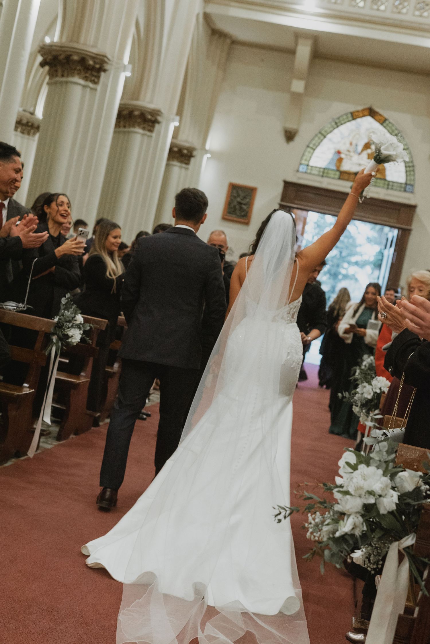 Bride and groom walk down church aisle after wedding ceremony, guests applauding, bride raising bouquet in celebration.