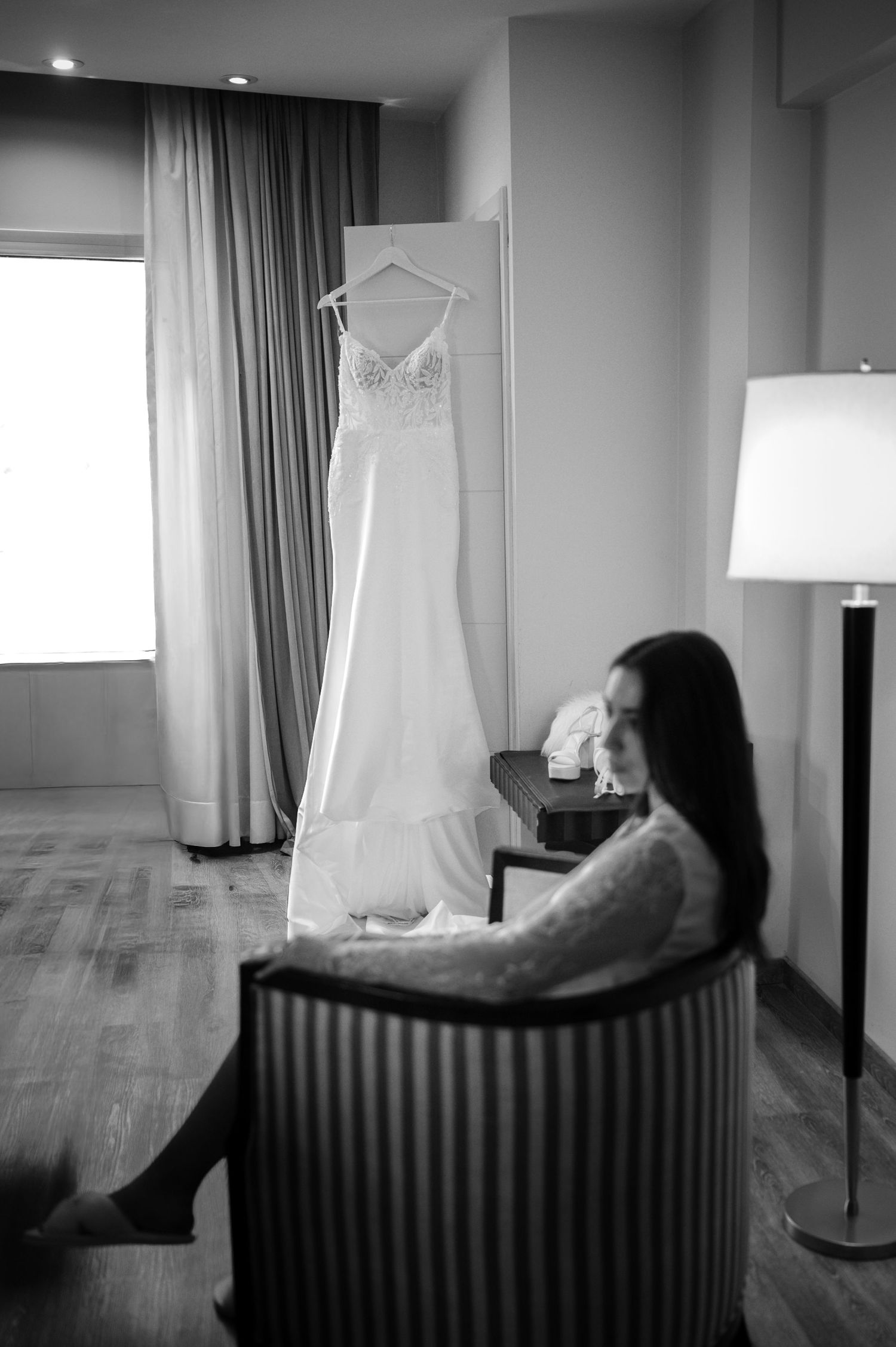 Black and white photo of bride sitting in chair gazing at wedding dress hanging near window in hotel room.
