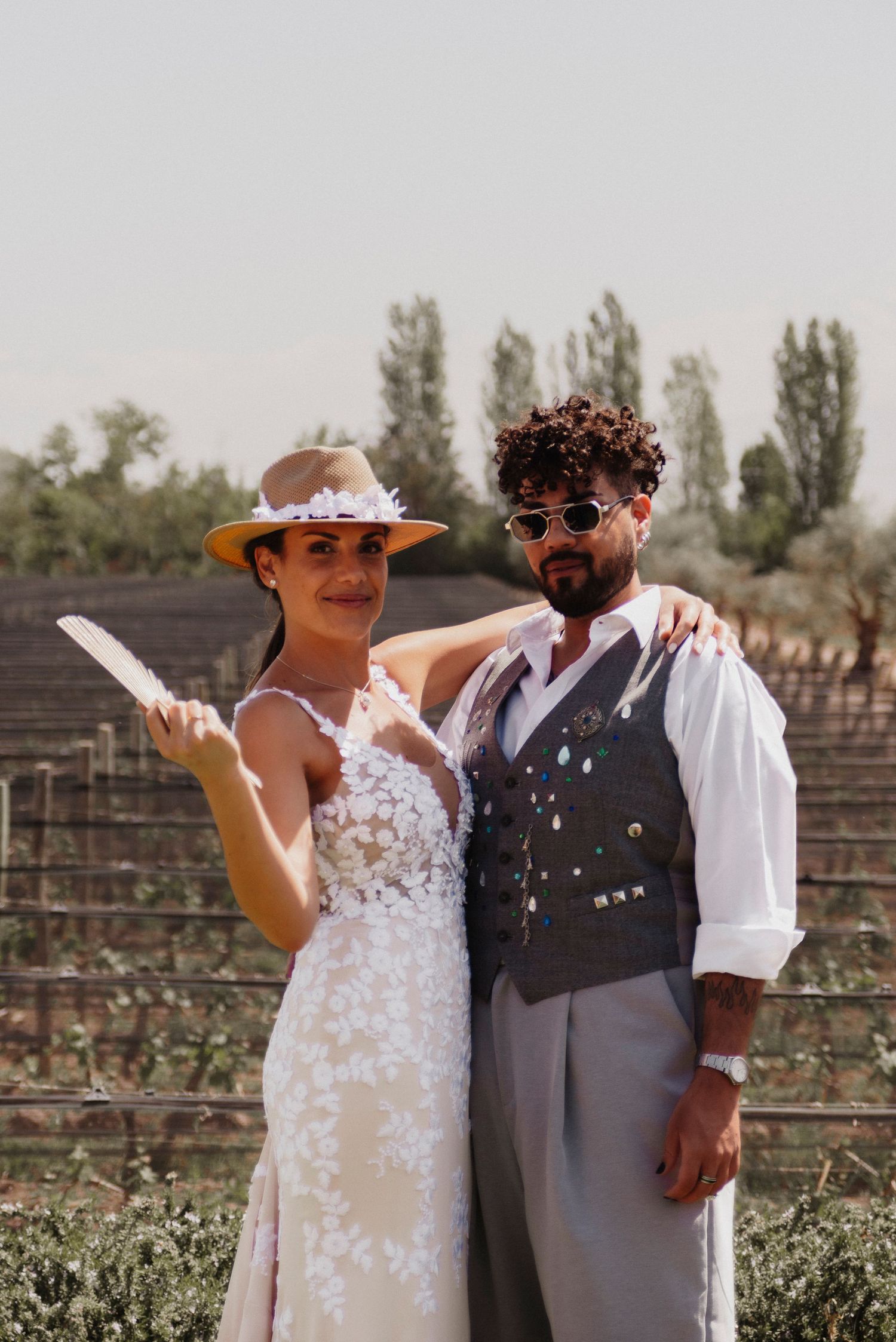 A couple poses together in a vineyard on their wedding day wearing a white dress and gray vest.