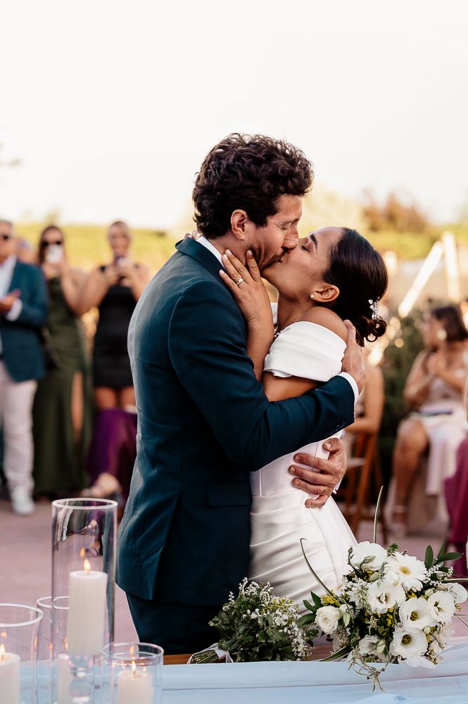 A romantic wedding kiss at an outdoor reception with white floral table decorations and sunset lighting.