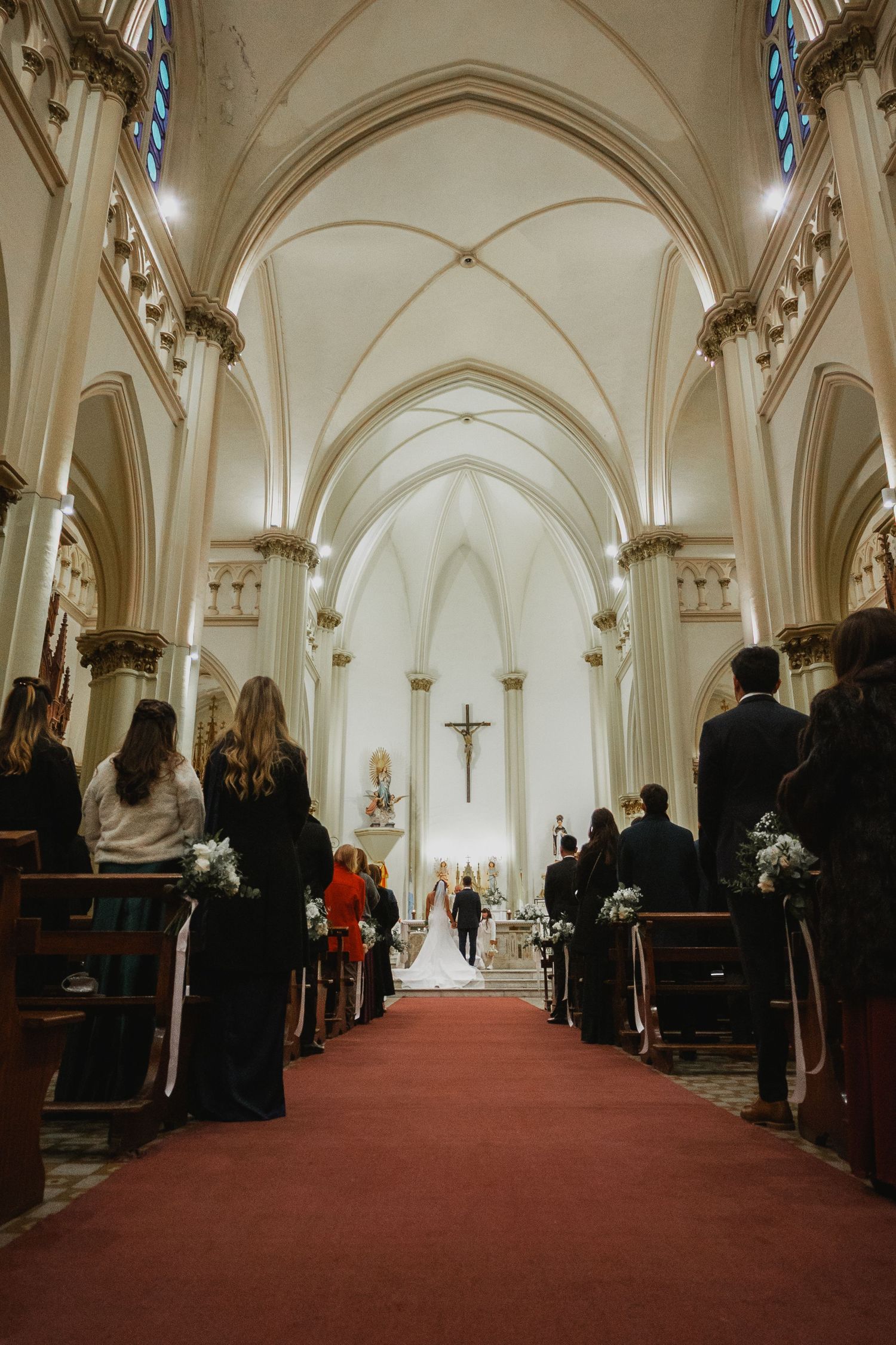 Wedding ceremony in a grand white Gothic church with red carpet aisle, guests seated in pews, couple at altar beneath crucifix.