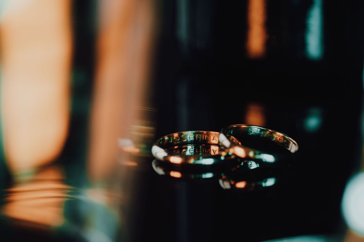 Dos anillos de boda de oro descansan sobre una superficie oscura y reflectante, con una cálida luz bokeh de fondo.