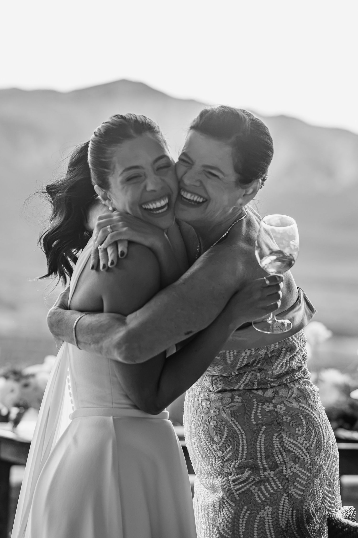 Two women share a joyful hug at an outdoor wedding, one holding a wine glass, smiling against a mountain backdrop.