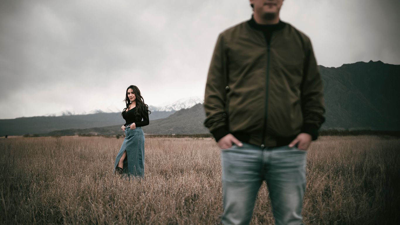 Two figures stand apart in a moody field against mountain backdrop during golden hour.