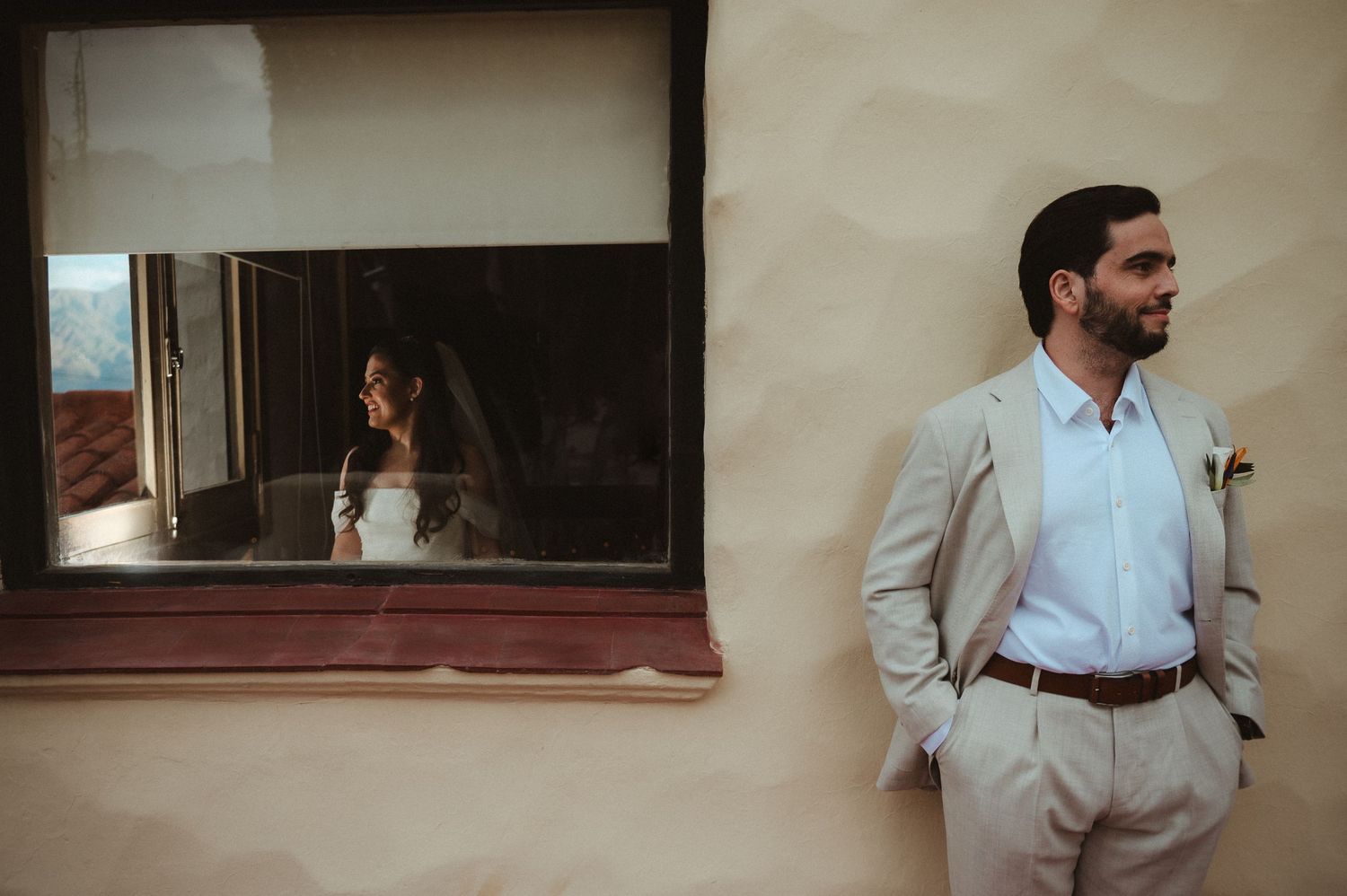 Stylish wedding couple poses separately with groom in beige suit against wall and bride visible through window reflection.