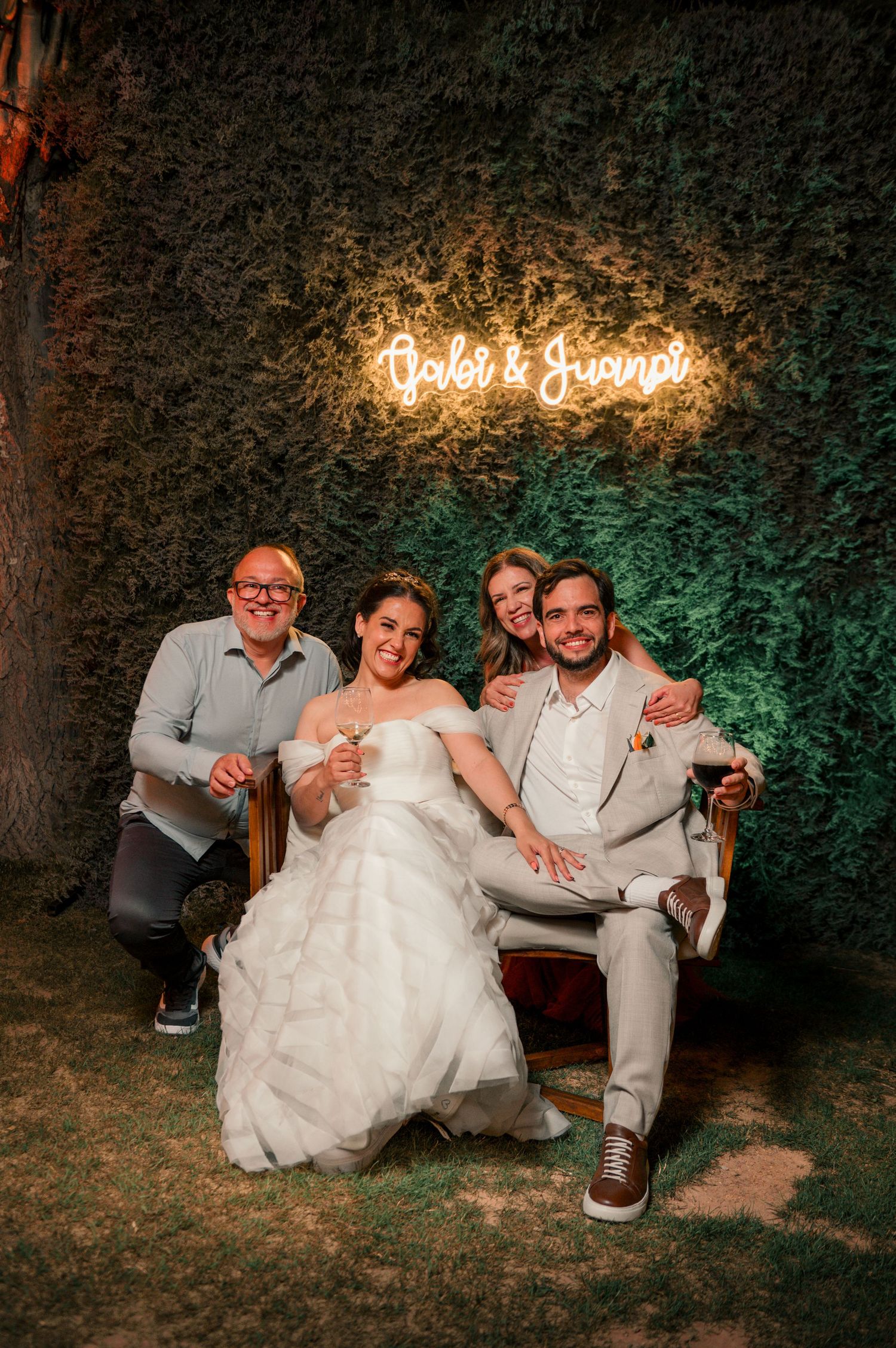 Wedding party members relax and laugh together on a bench against a dark garden backdrop with illuminated signage.