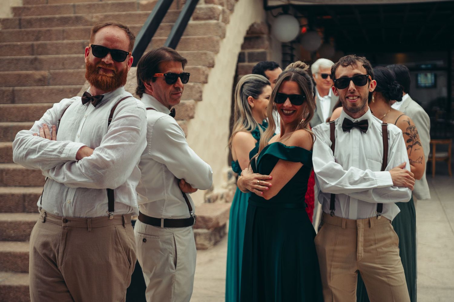 Wedding party members in sunglasses and formal attire pose together in a casual outdoor setting.