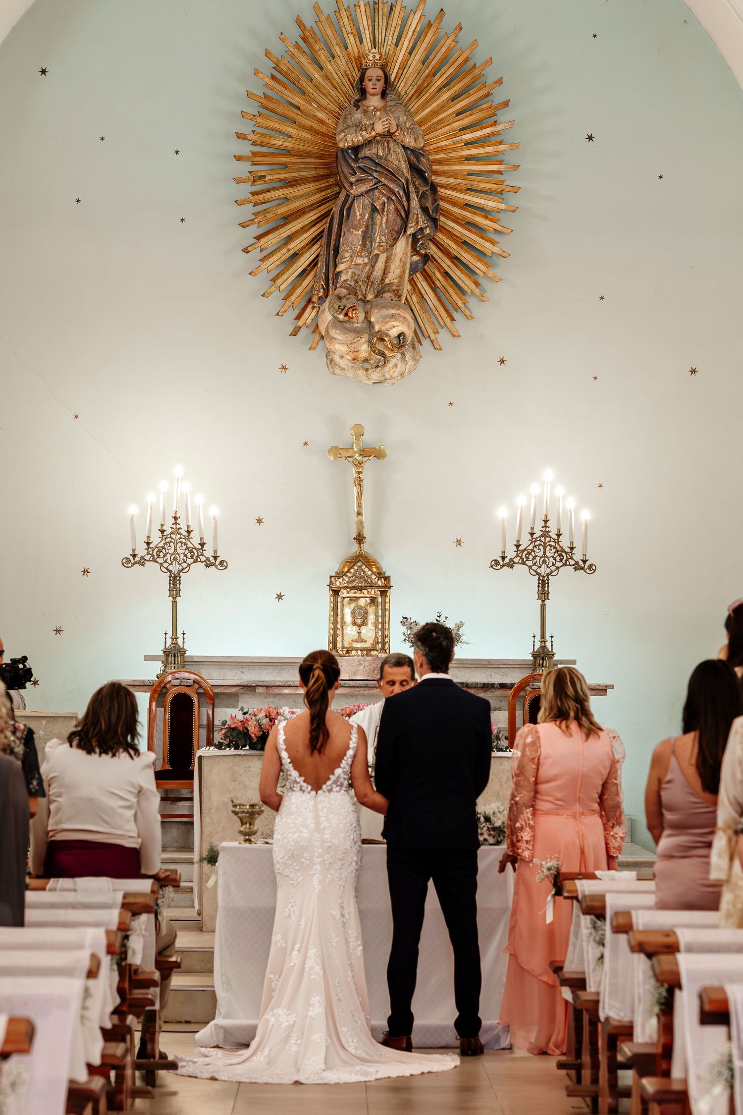 Wedding guests stand in a traditional church sanctuary with ornate religious decor and a sunburst design above the altar.