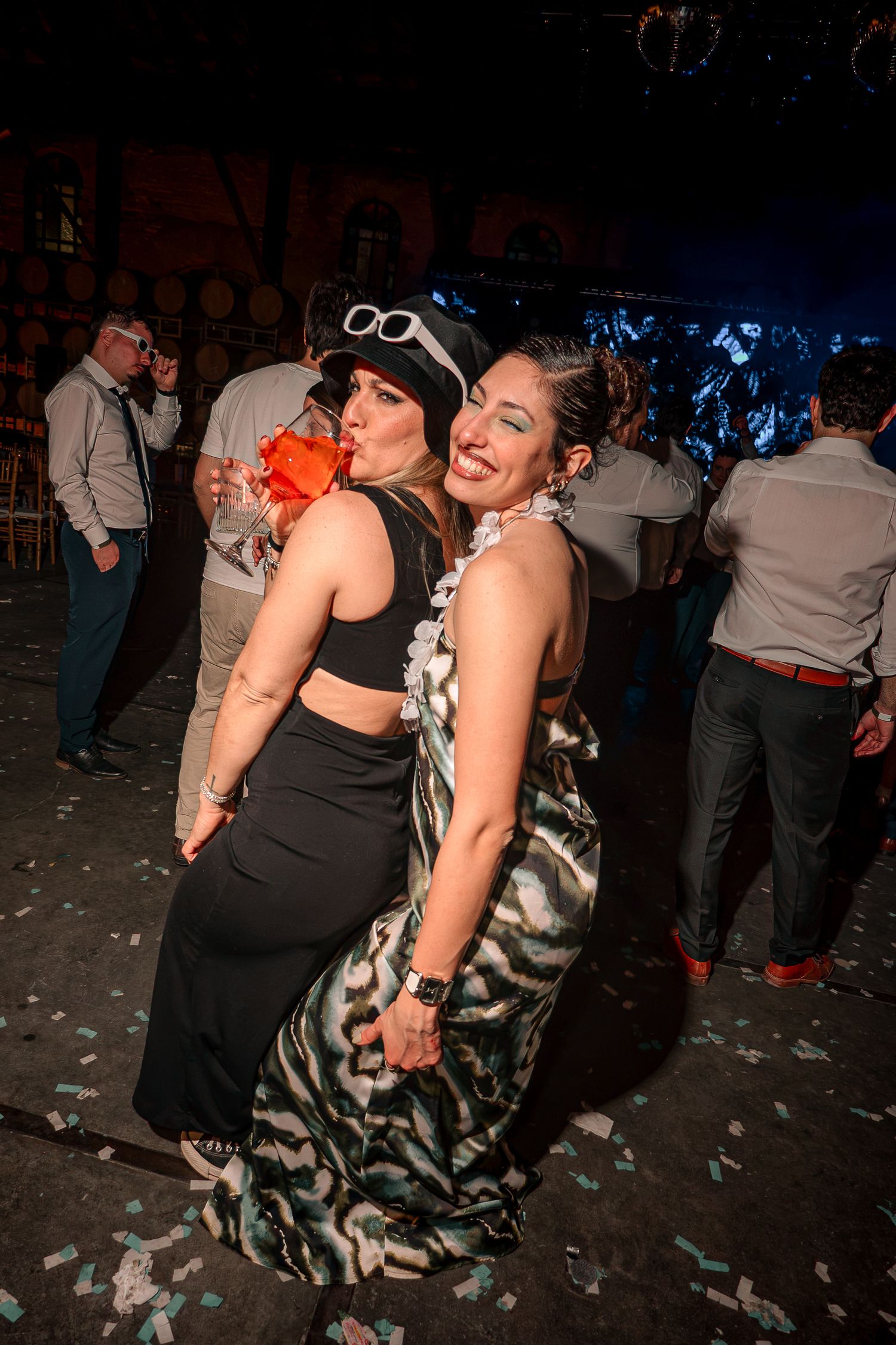 Two women in festive costumes pose together on a confetti-covered dance floor at a lively party.