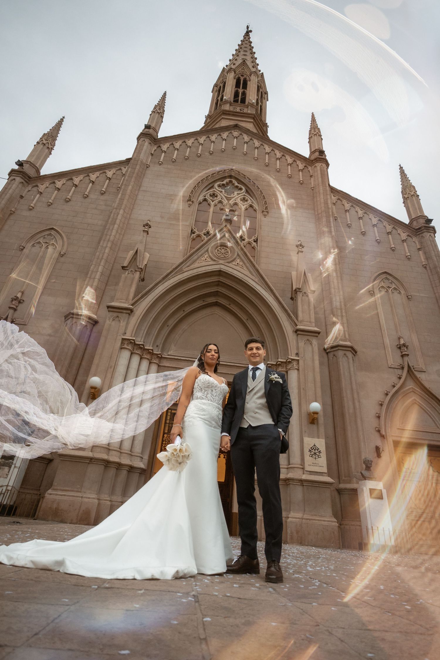 Bride and groom holding hands before gothic stone church, veil flowing dramatically in wind during outdoor wedding portrait.