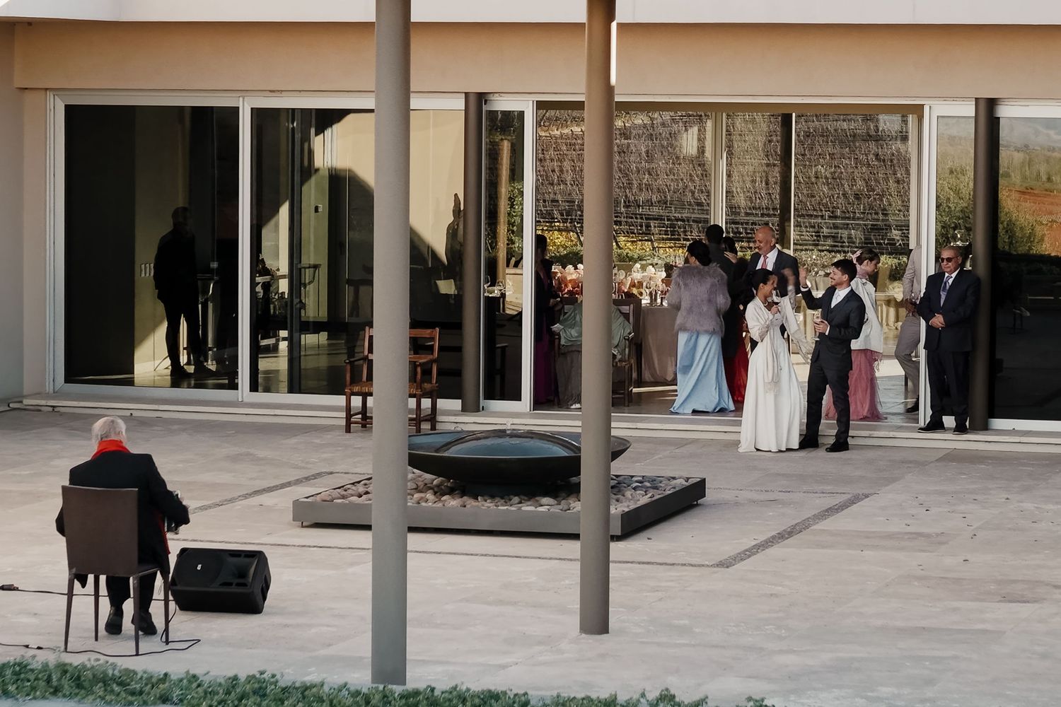 Outdoor wedding ceremony on a modern patio with guests gathered, a seated musician, and large glass windows in the background.