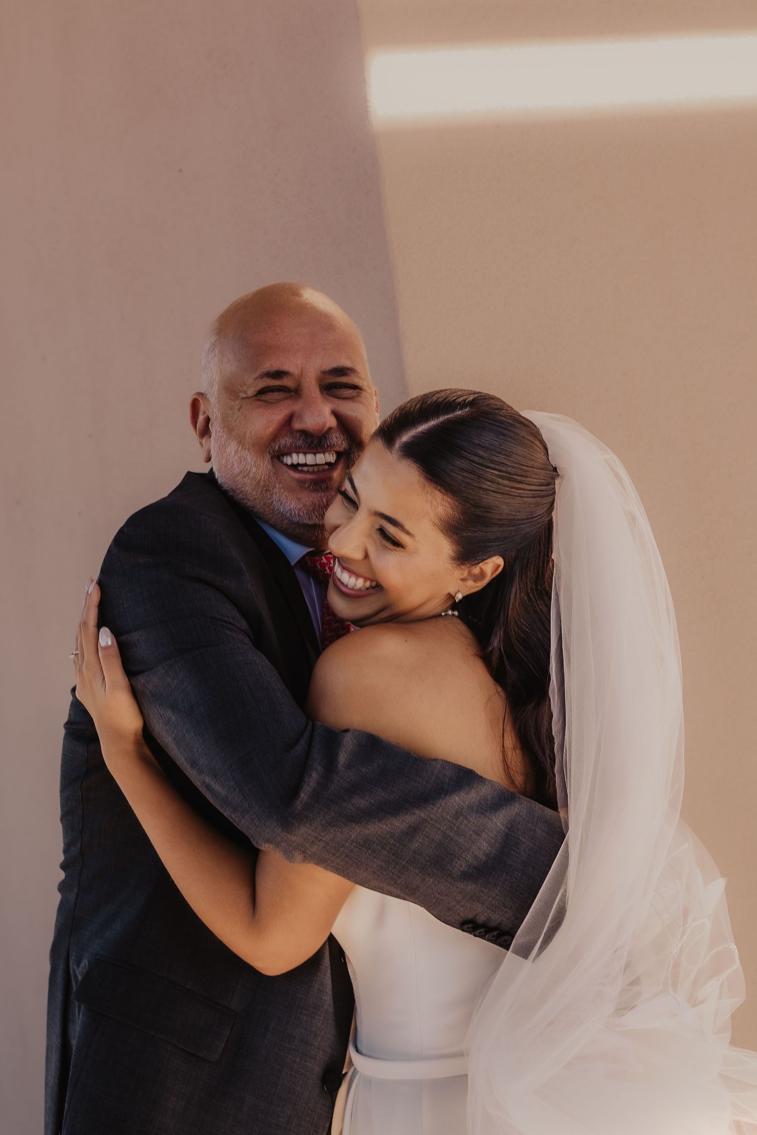 A bride in a white gown and veil shares a joyful hug with an older man in a suit on her wedding day.