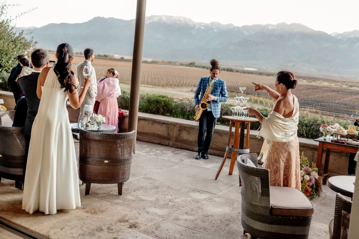 Outdoor wedding reception on a vineyard terrace with guests, a saxophonist in blue, and mountain views in the background.