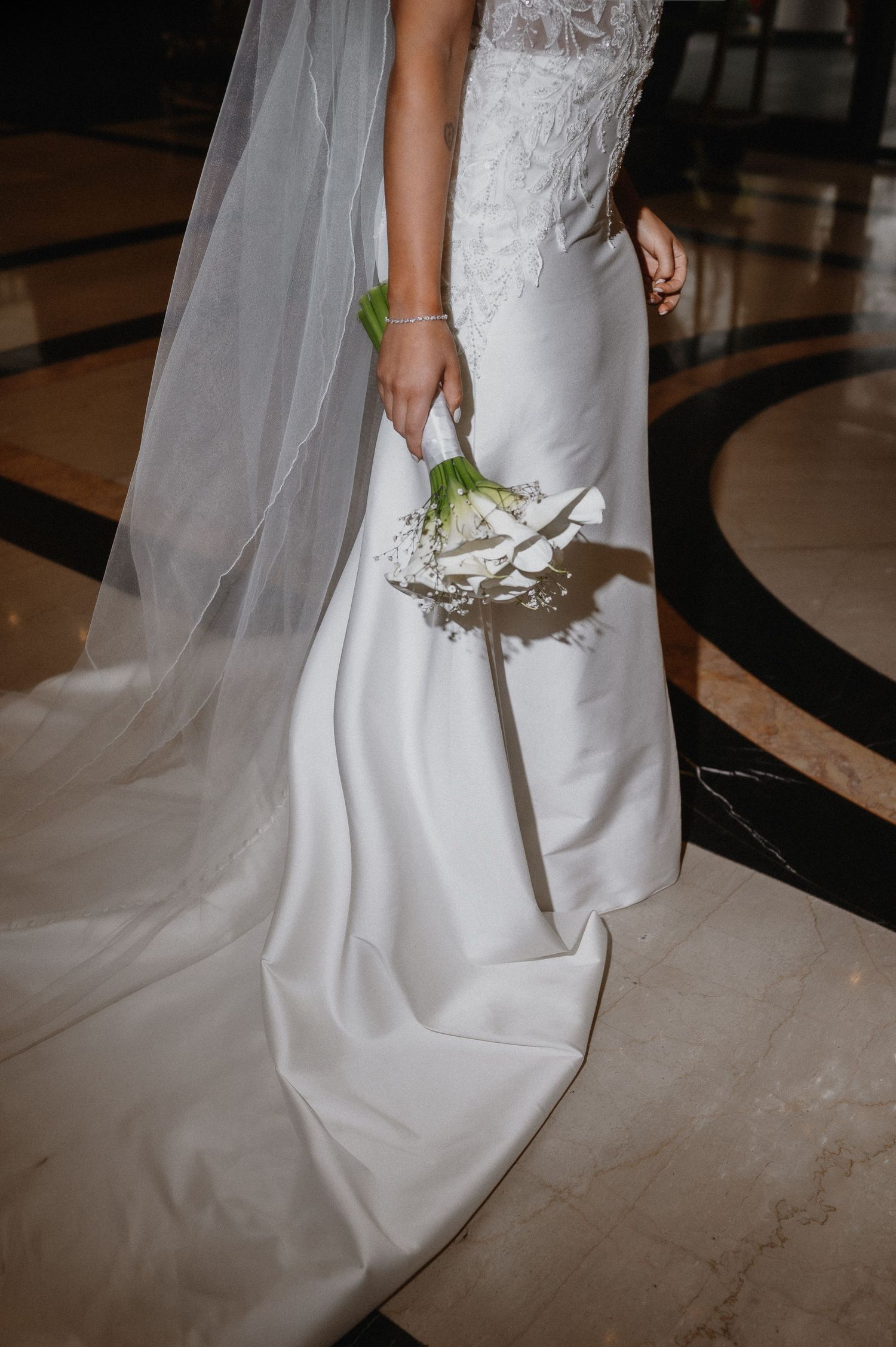 Bride in white mermaid wedding gown with lace bodice, flowing veil, and white rose bouquet on marble floor.
