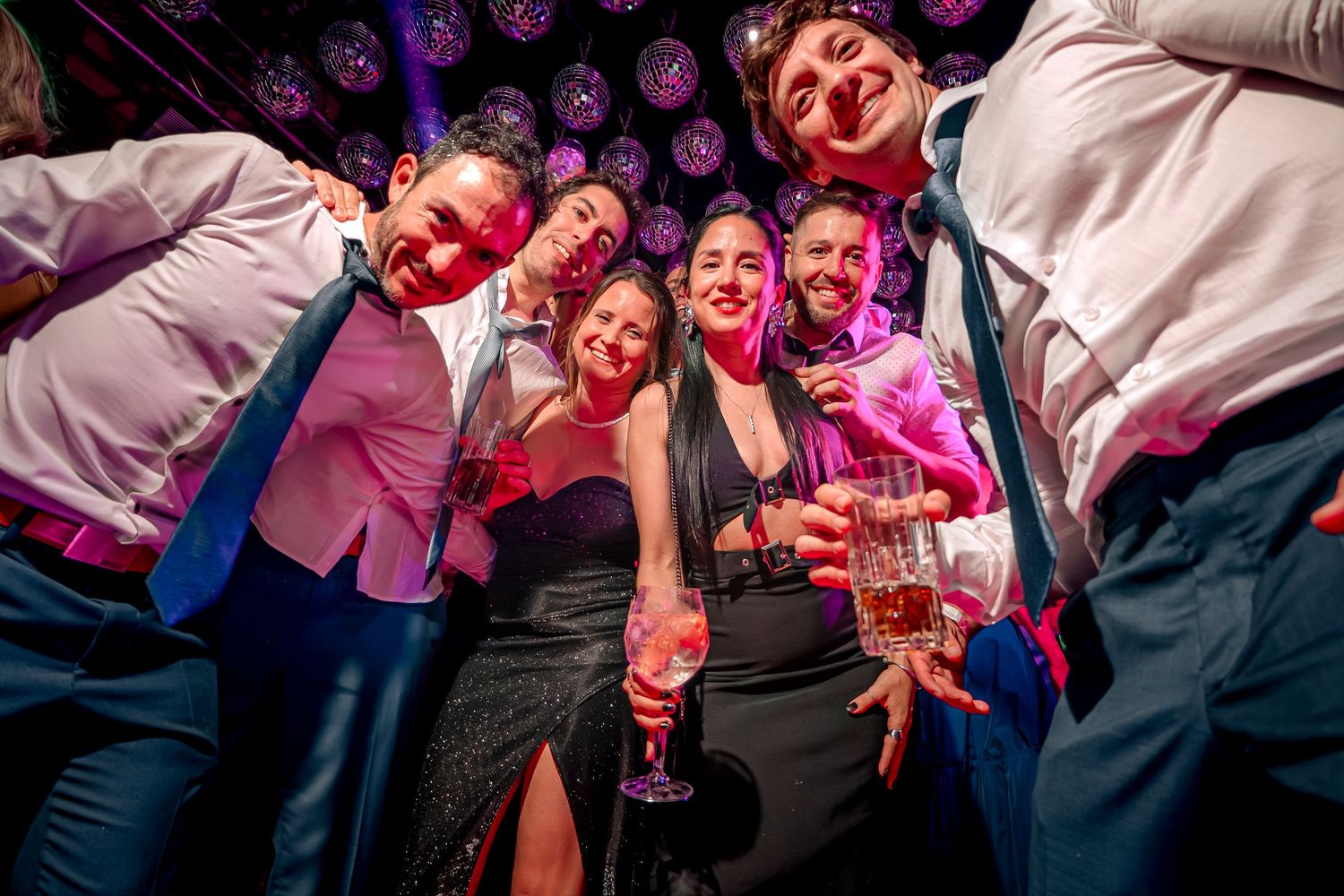 A lively crowd dances and celebrates together at a wedding reception party under pink and purple lighting.