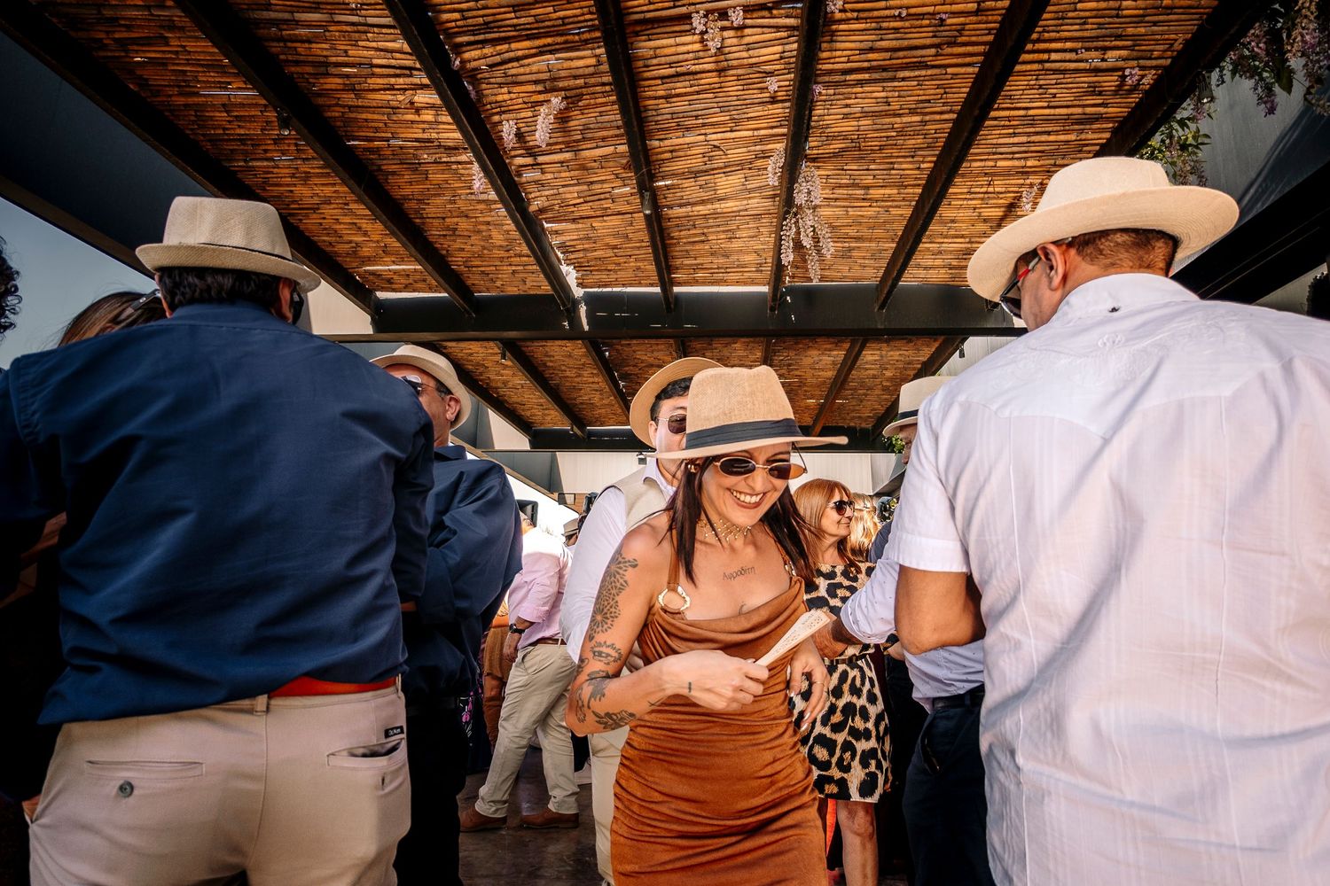 A group of people in casual attire and sun hats gather under a rustic wooden ceiling at an outdoor event.