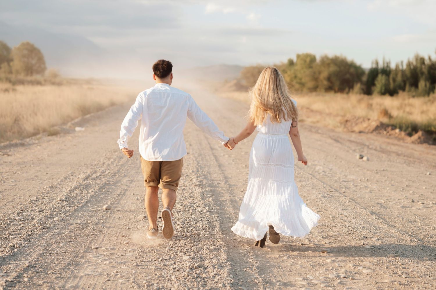 Couple in white and beige outfits walking hand in hand down a dusty country road at sunset.