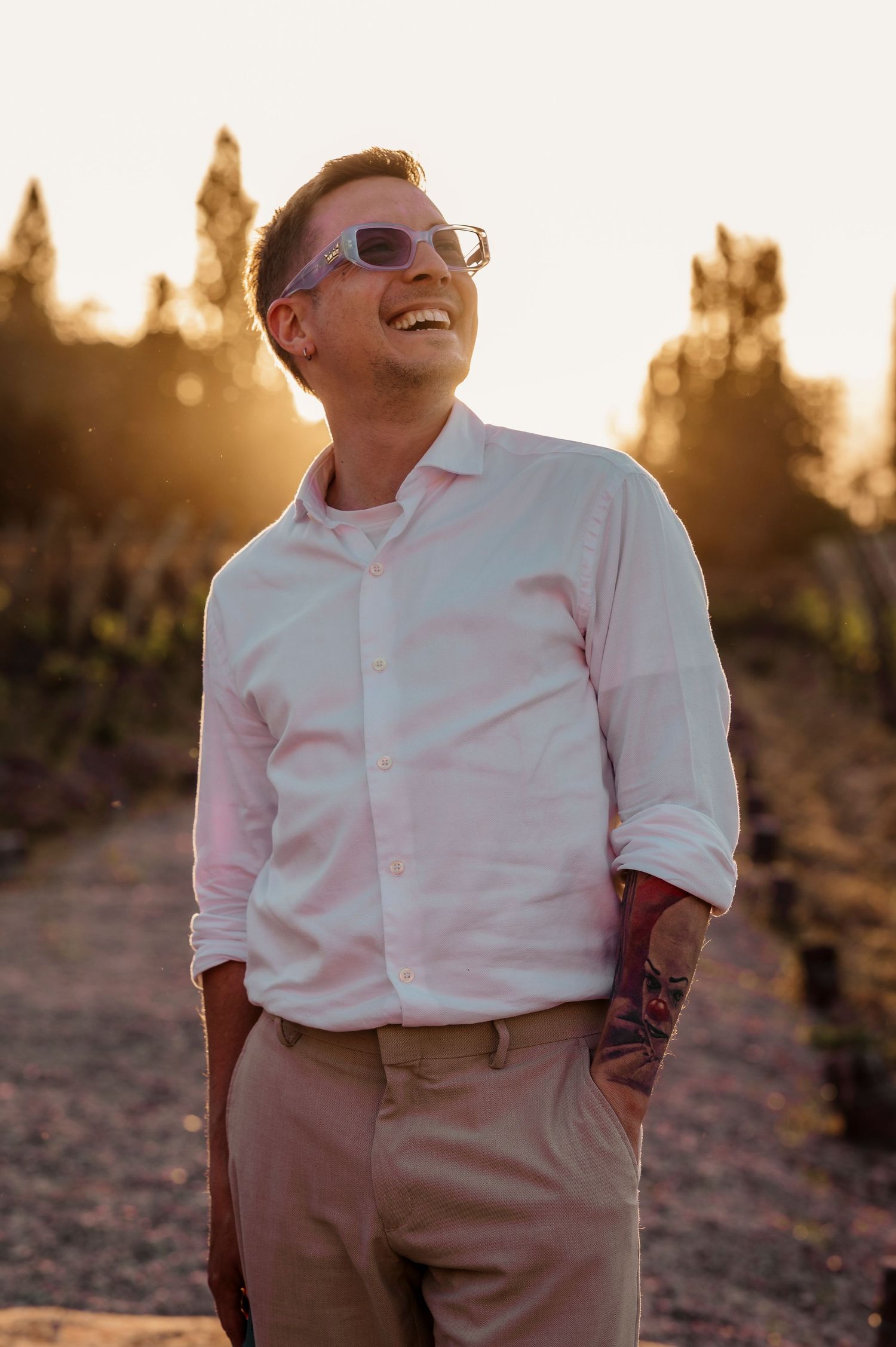A person in sunglasses wearing a white button-down shirt and khaki pants poses at sunset in a desert landscape.