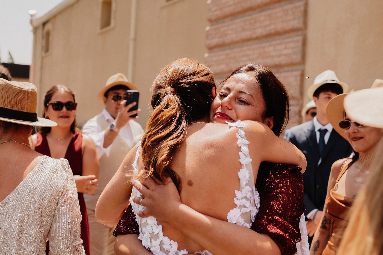 Two people share an emotional embrace at an outdoor celebration while guests look on in the sunlight.