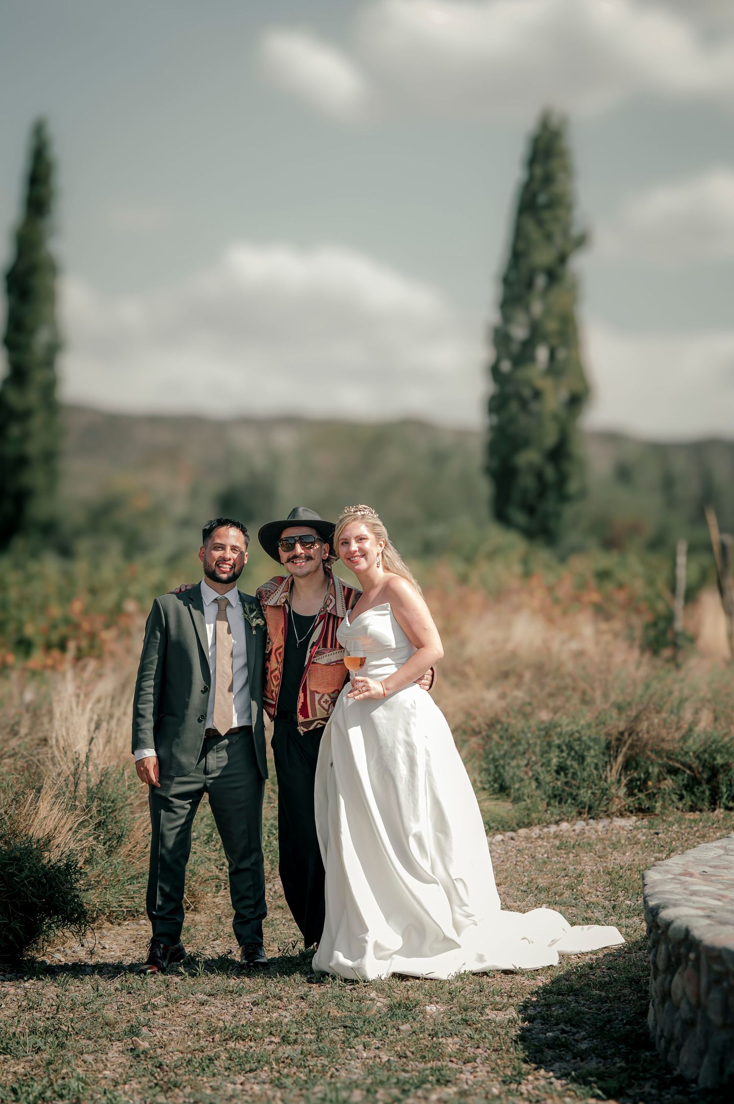 Wedding party poses together in a rustic outdoor setting with tall grass and cypress trees in the background.