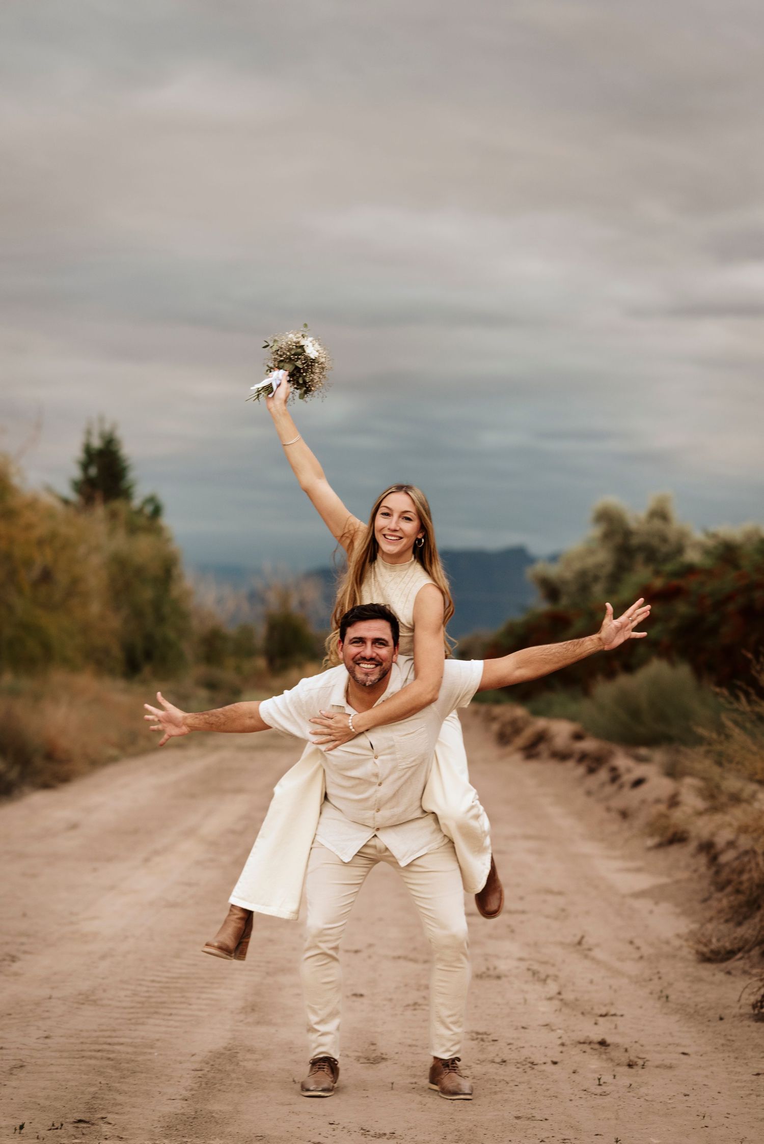 Playful wedding couple poses joyfully on a dirt road with mountains in the background during golden hour.