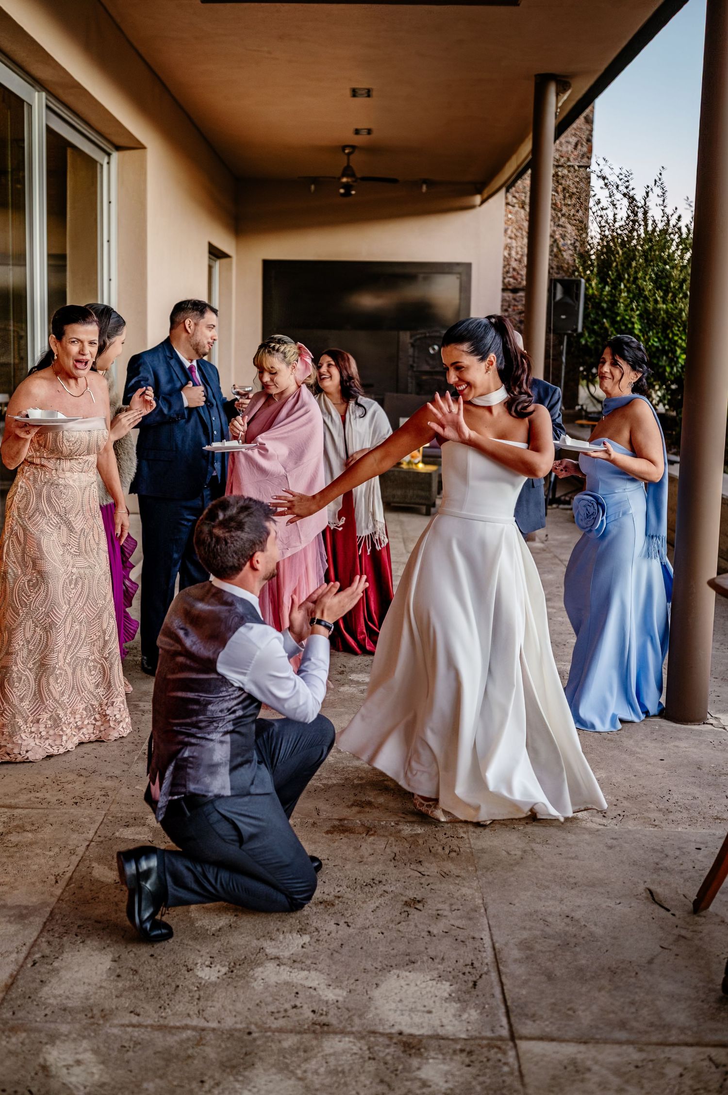 Groom kneeling before bride in white dress during wedding reception dance, surrounded by guests in formal attire outdoors.