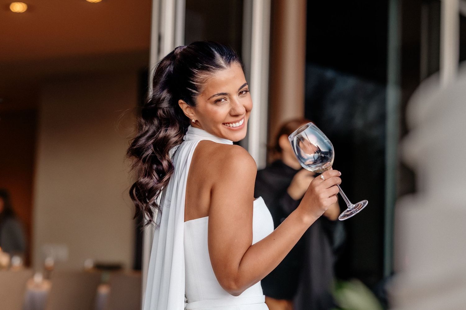 Smiling bride in white dress holding a wine glass, with long dark wavy hair at an elegant event.
