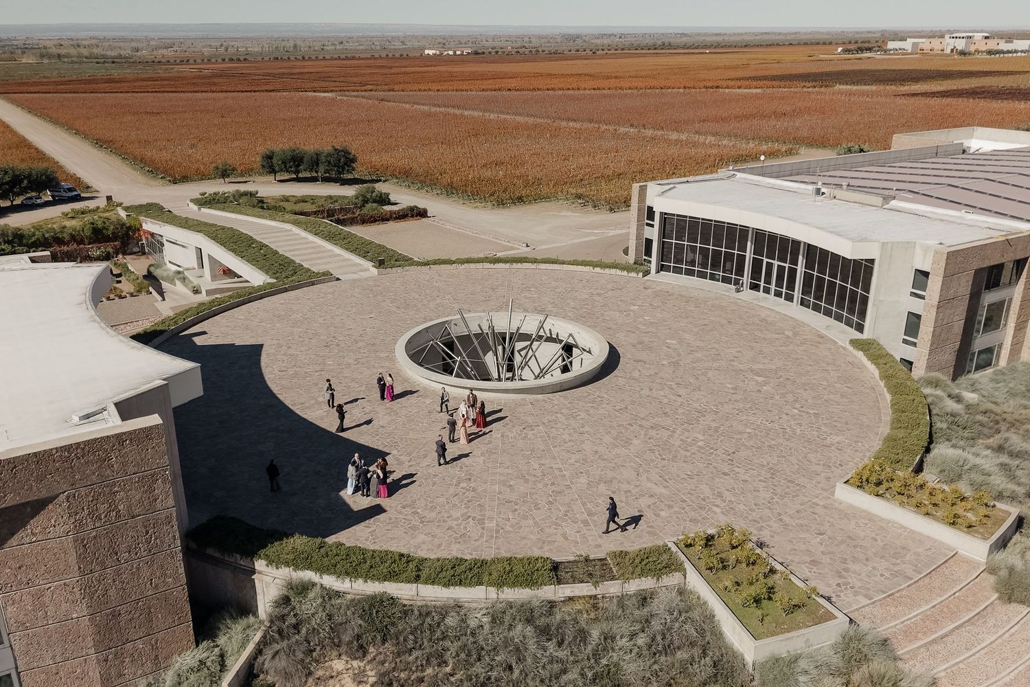 Aerial view of a modern campus courtyard with circular paved plaza, central sculpture, and vast farmland backdrop.fotógrafos de boda Mendoza