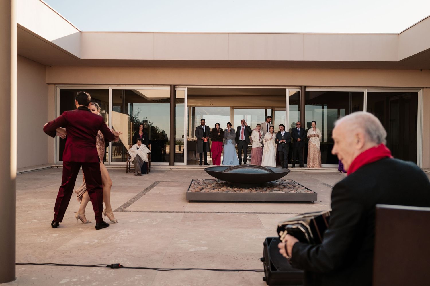 Wedding party poses on a modern patio with fire pit while couple dances and accordion player performs at dusk.