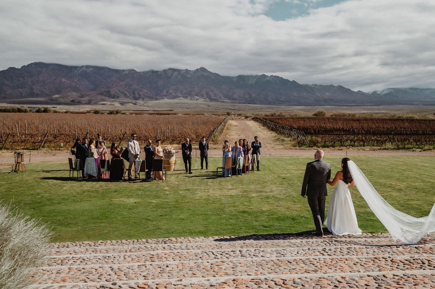 Outdoor vineyard wedding ceremony with bride and groom walking toward guests, mountain backdrop, South Africa winery.