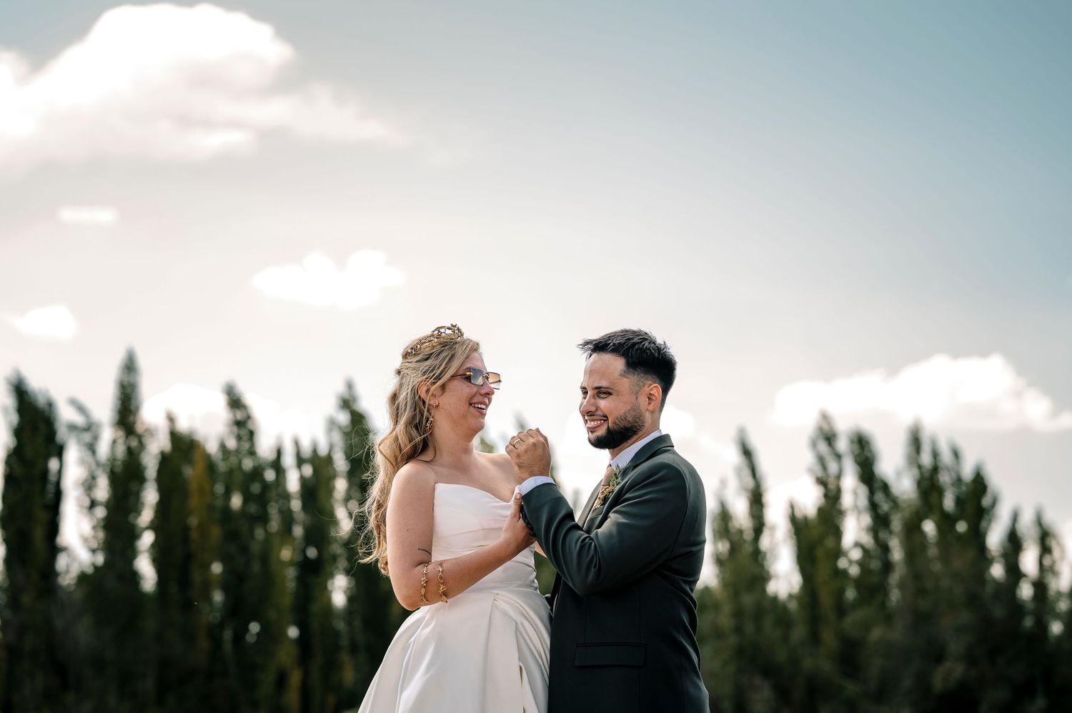 A couple shares romantic moments in a wedding photoshoot against a backdrop of tall trees and blue sky.