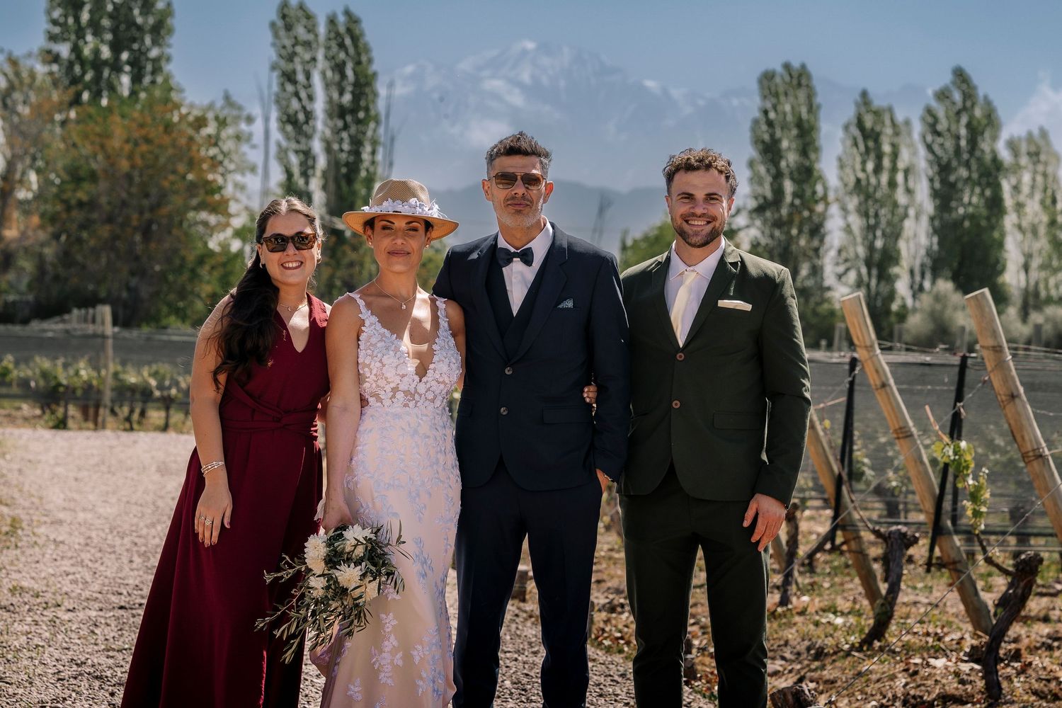 Wedding party poses together outdoors with tall trees and blue sky in background on sunny day.