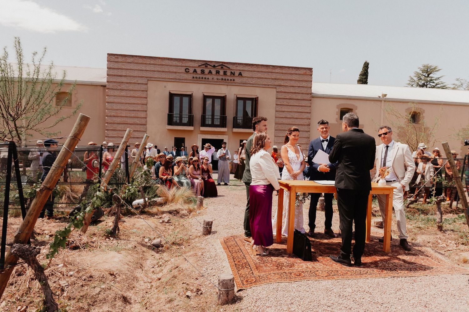 Outdoor desert wedding ceremony with guests gathered in front of a pink stucco building surrounded by cacti and desert plants.