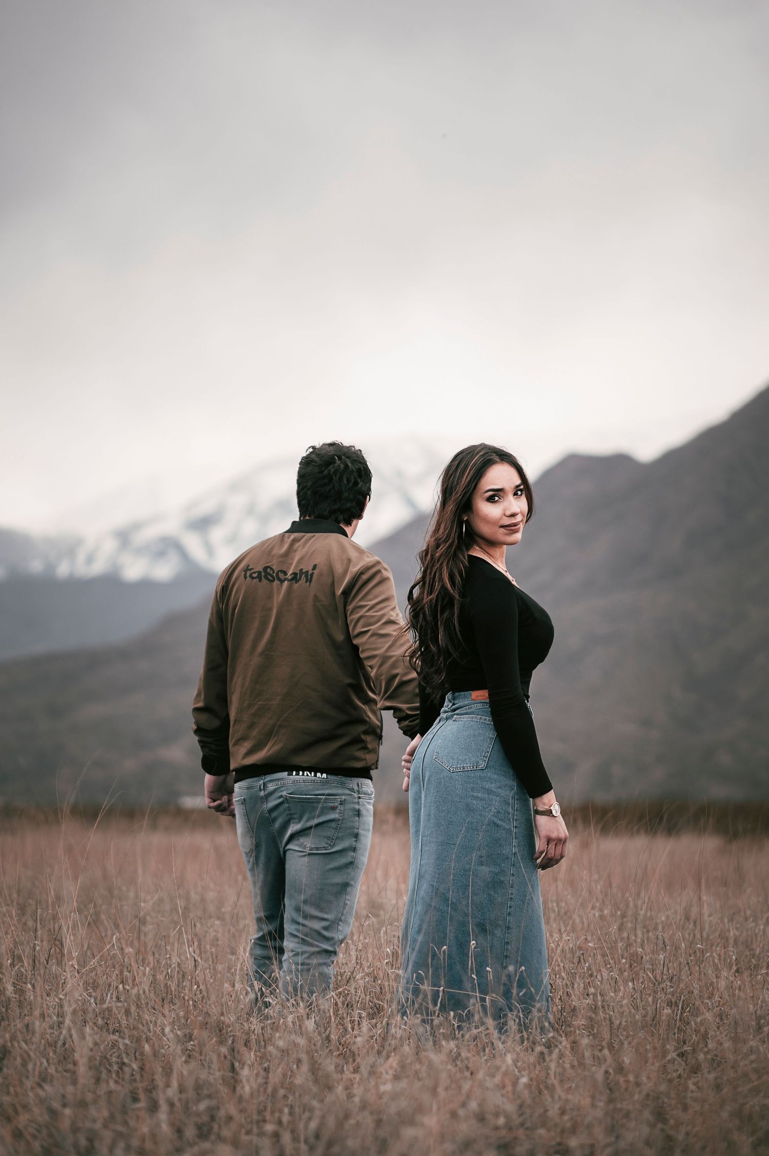 Couple standing in field with snow-capped mountains visible in background.