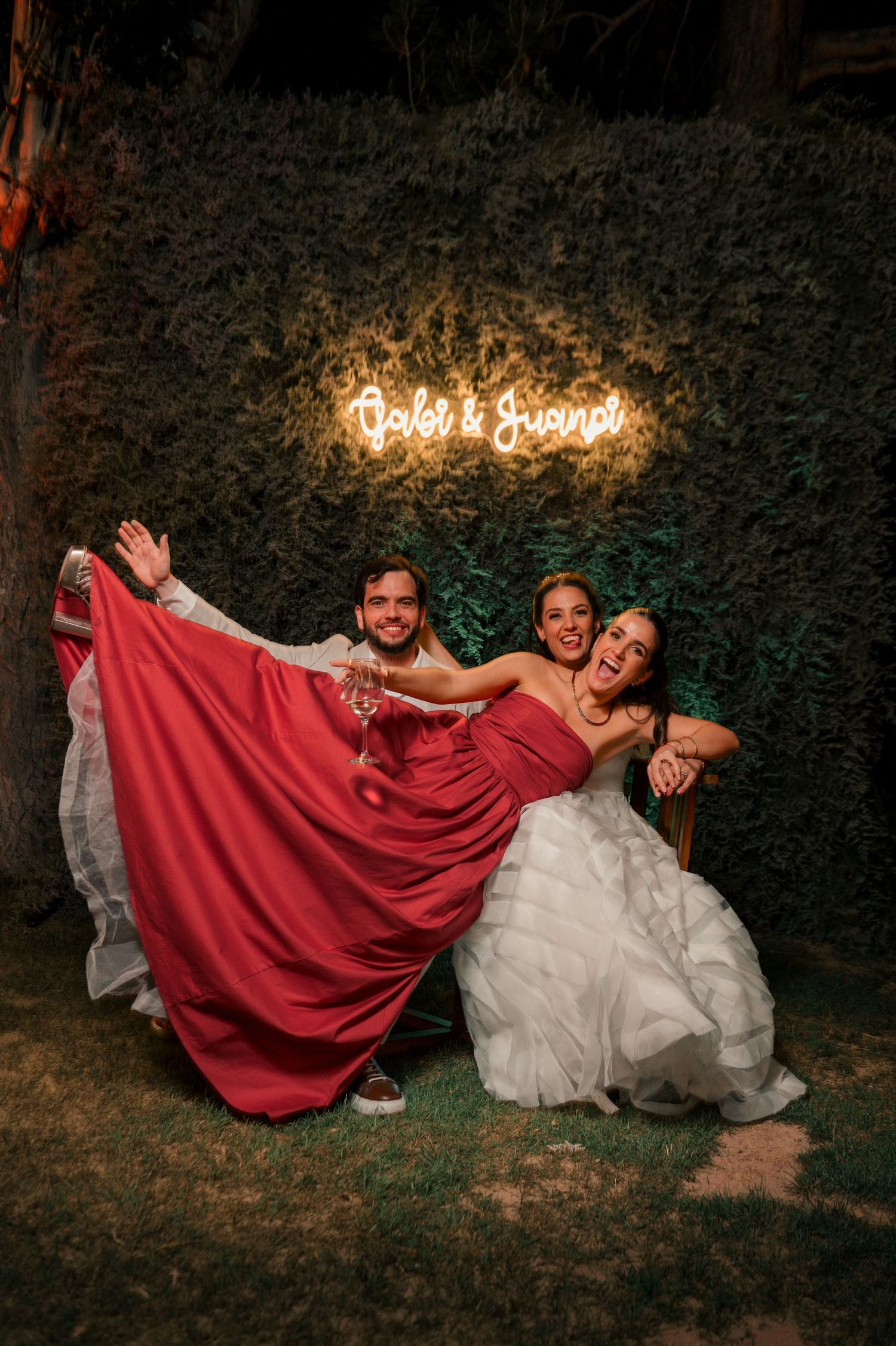 Three people in formal attire pose playfully under illuminated 'God's Grace' sign against a hedge wall at night.