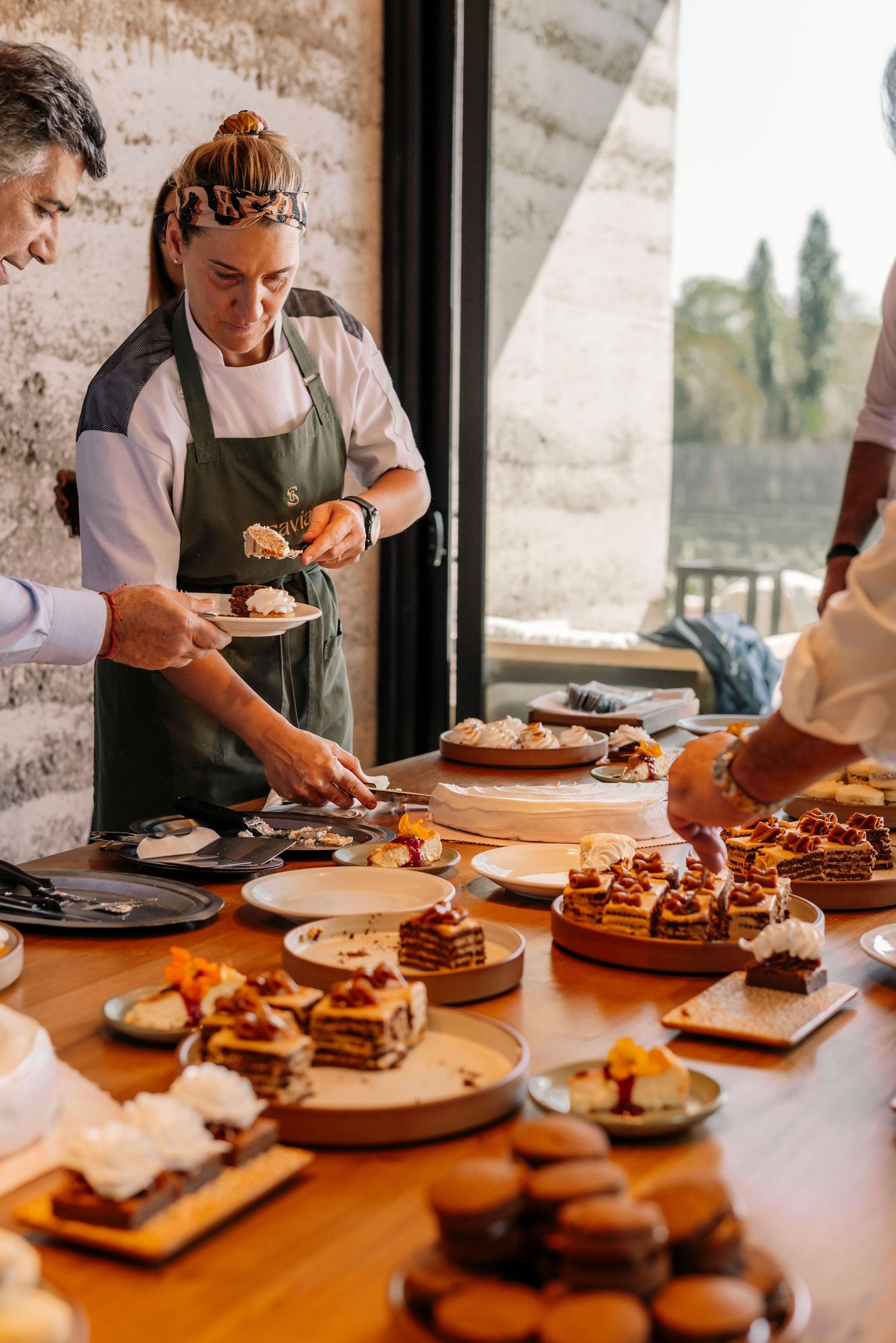 Chefs in aprons prepare and plate desserts on wooden boards at a rustic restaurant counter in warm lighting.