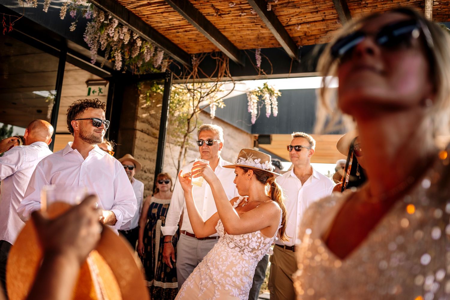 Wedding guests dancing and celebrating at an outdoor reception under wooden beams and string lights at sunset.