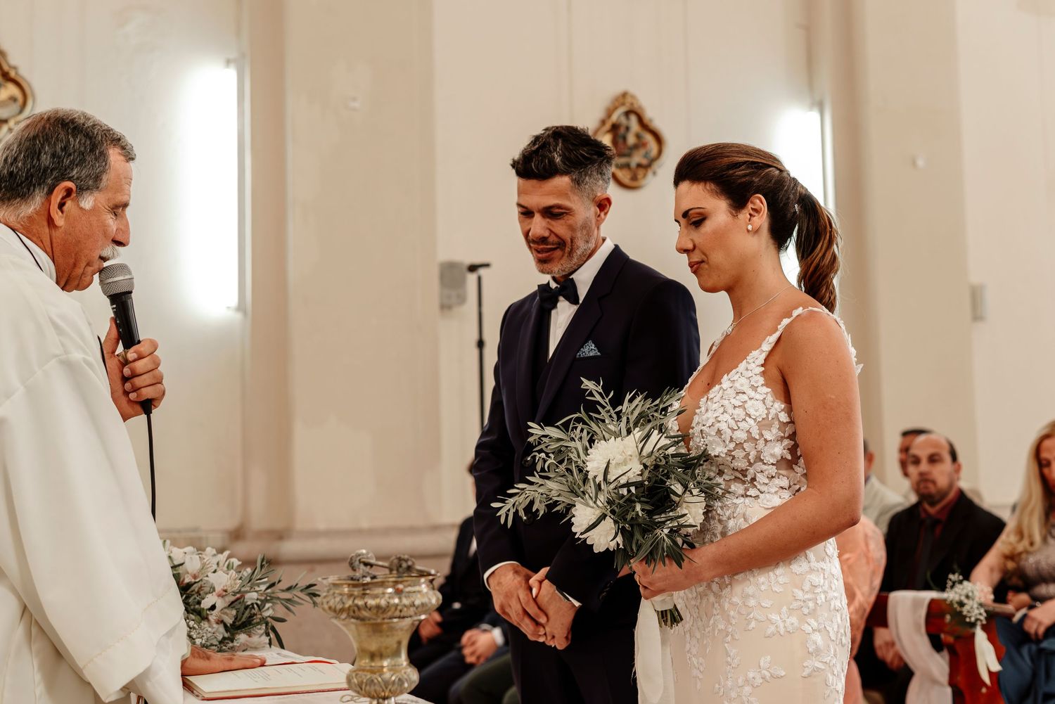 Wedding party gathers in an elegant white church interior during a traditional ceremony celebration.
