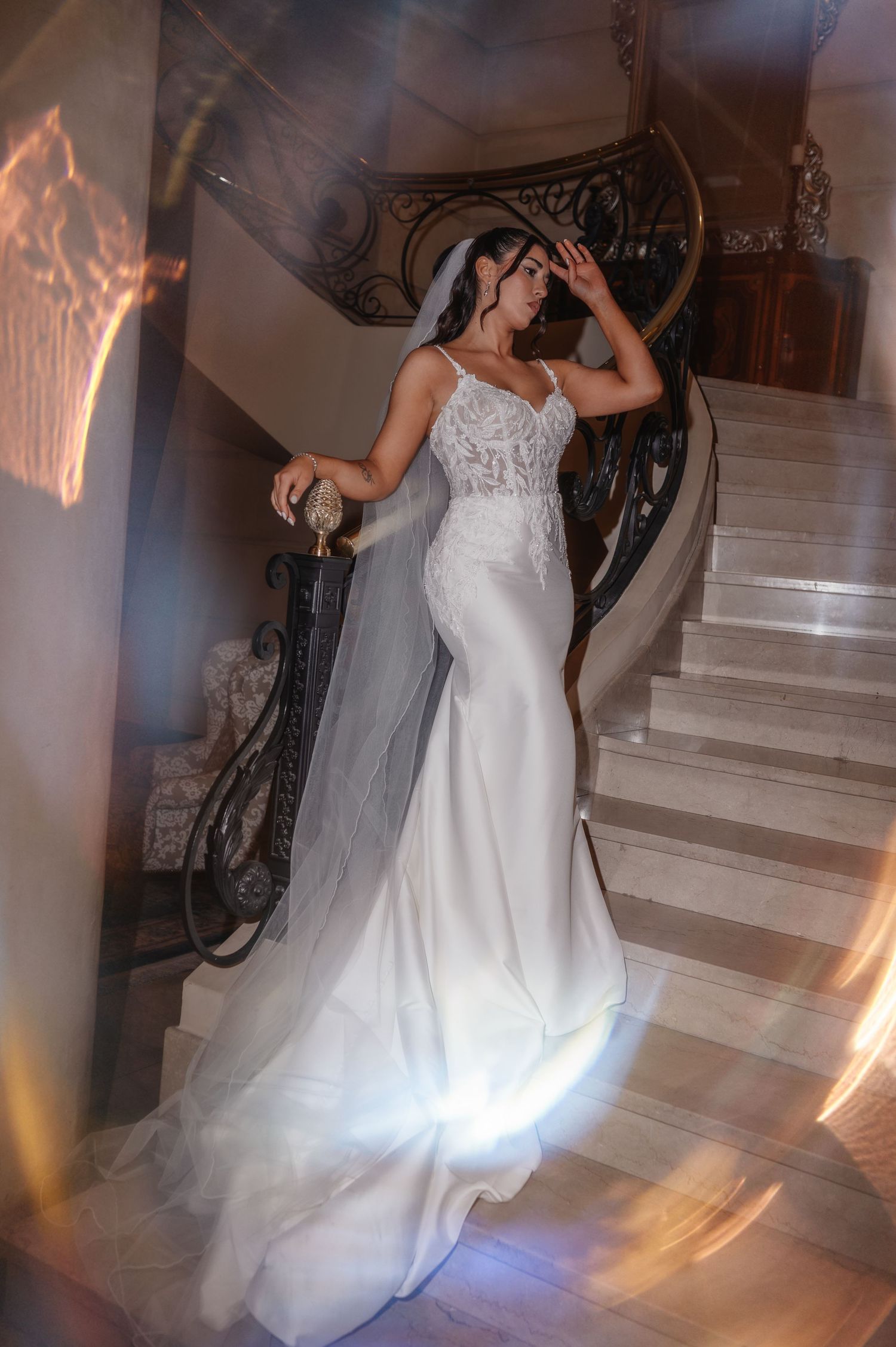 Bride in a fitted lace mermaid wedding gown with veil poses gracefully on an elegant marble staircase.