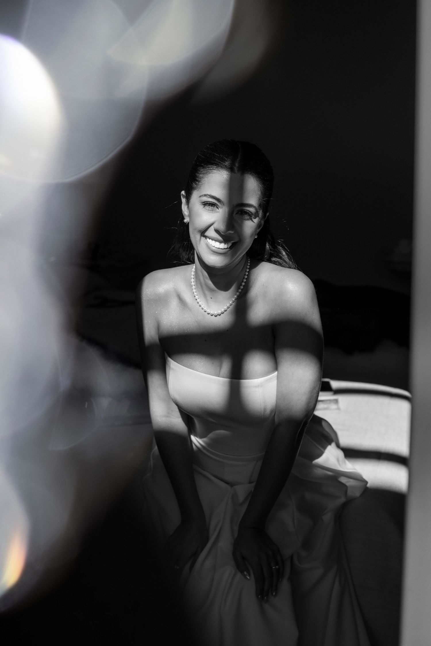 Black and white photo of a smiling woman in a strapless dress and pearl necklace, framed by soft bokeh light. fotógrafos de boda Mendoza