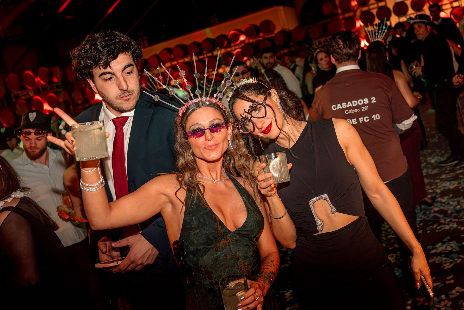 Three friends pose with drinks at a festive party, wearing fun accessories under colorful lights.