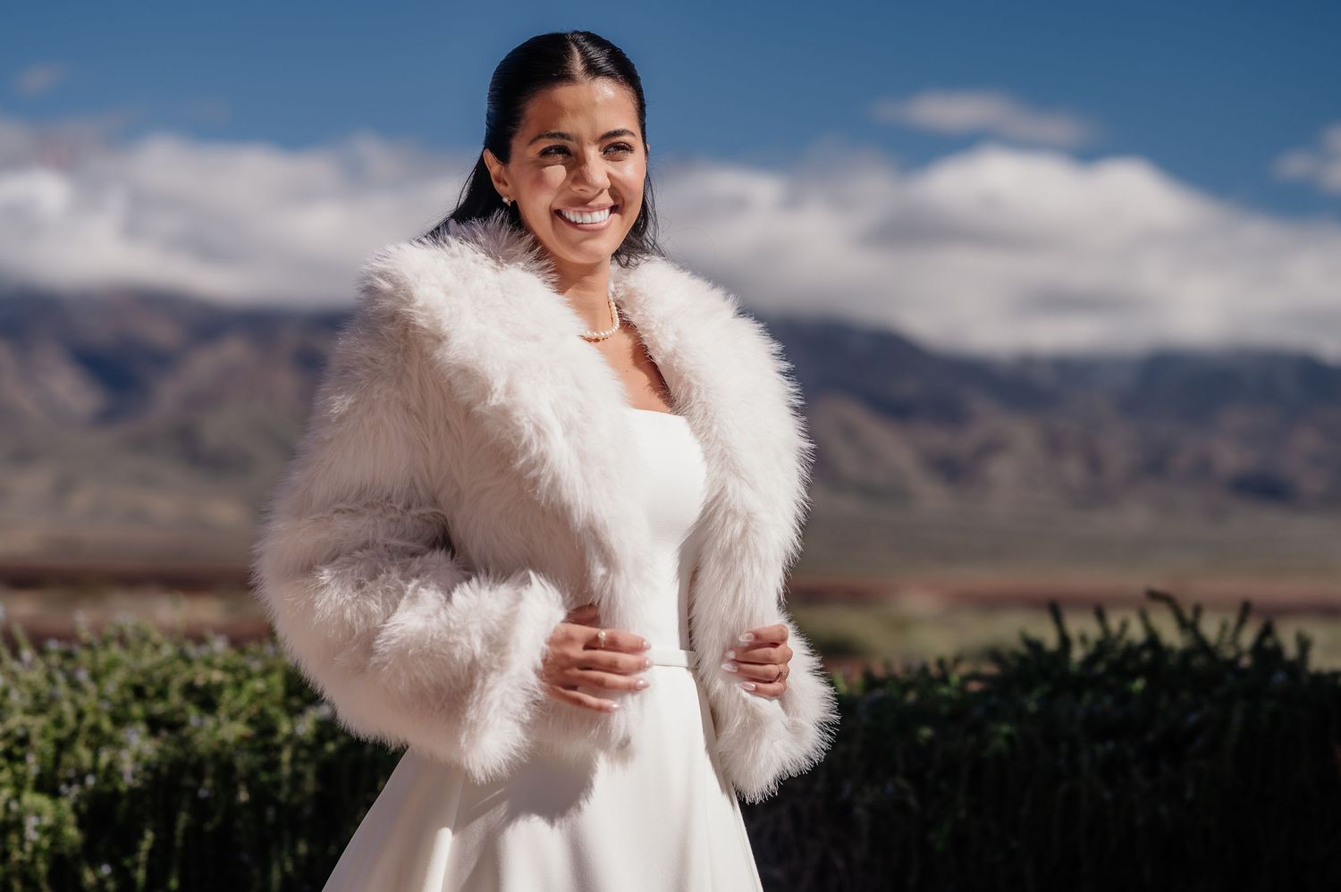 Bride smiling in white wedding dress and fluffy fur wrap jacket with mountain landscape backdrop.