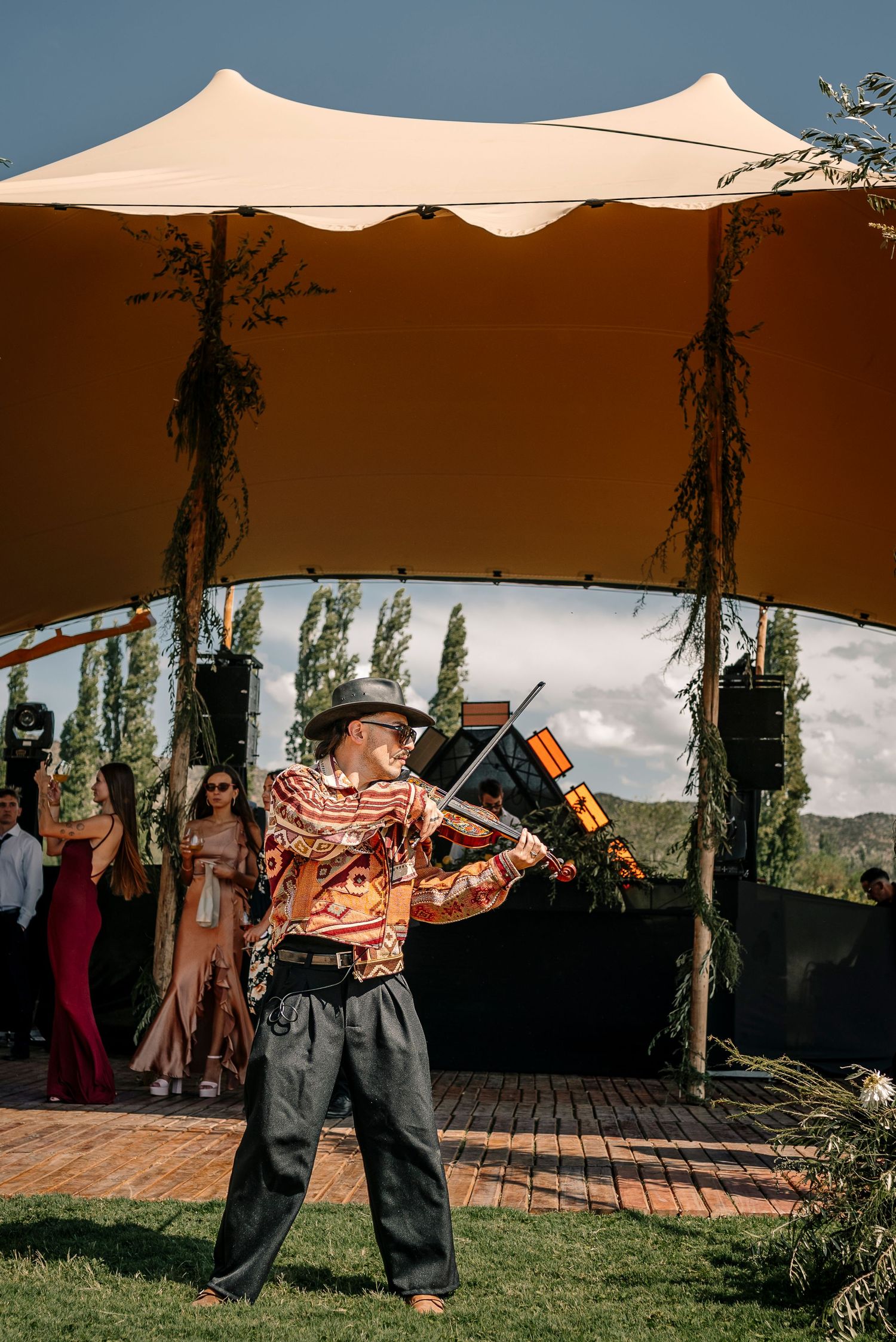A person in a hat and loose pants dances outdoors under a beige canopy tent on a sunny day.