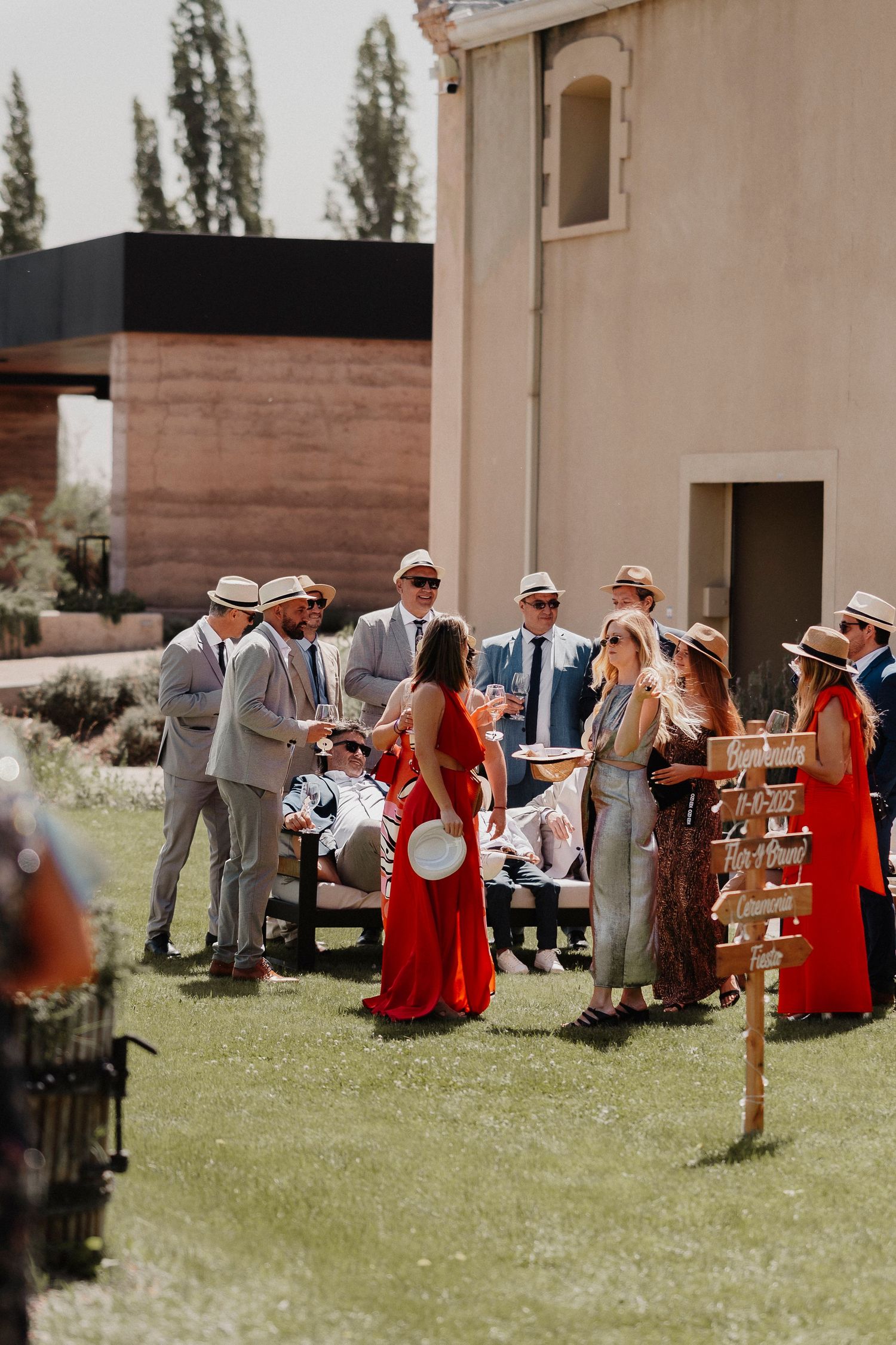 Wedding party in gray suits and red dresses gather on a sunny lawn during an outdoor celebration.