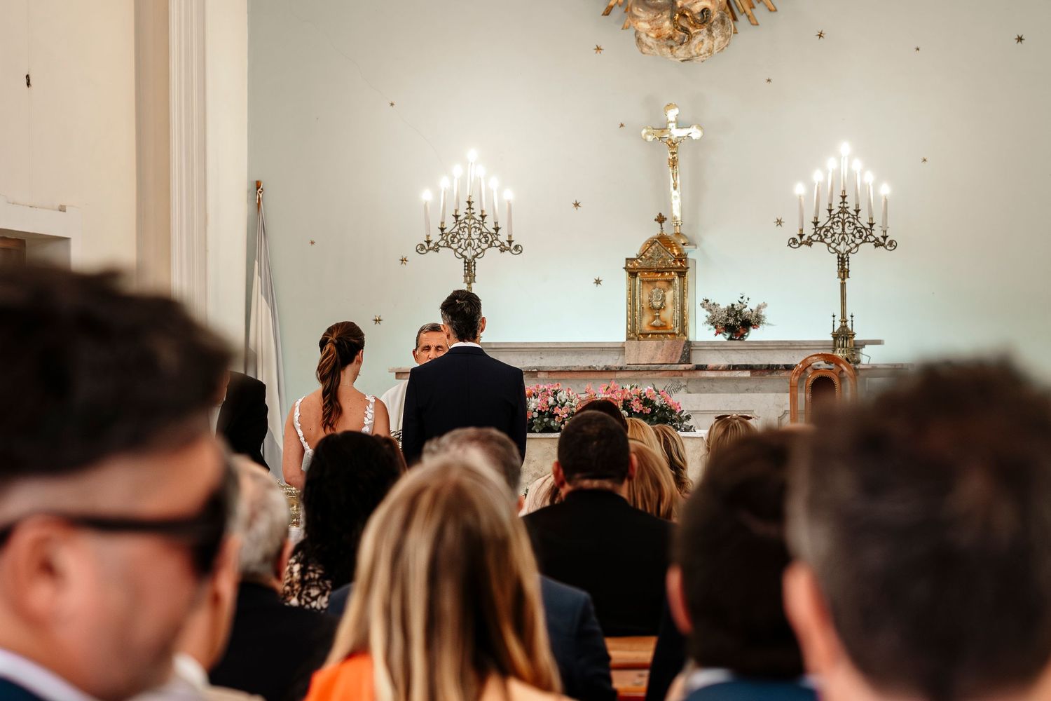 Wedding guests seated in church pews during ceremony with chandeliers hanging from ornate ceiling.