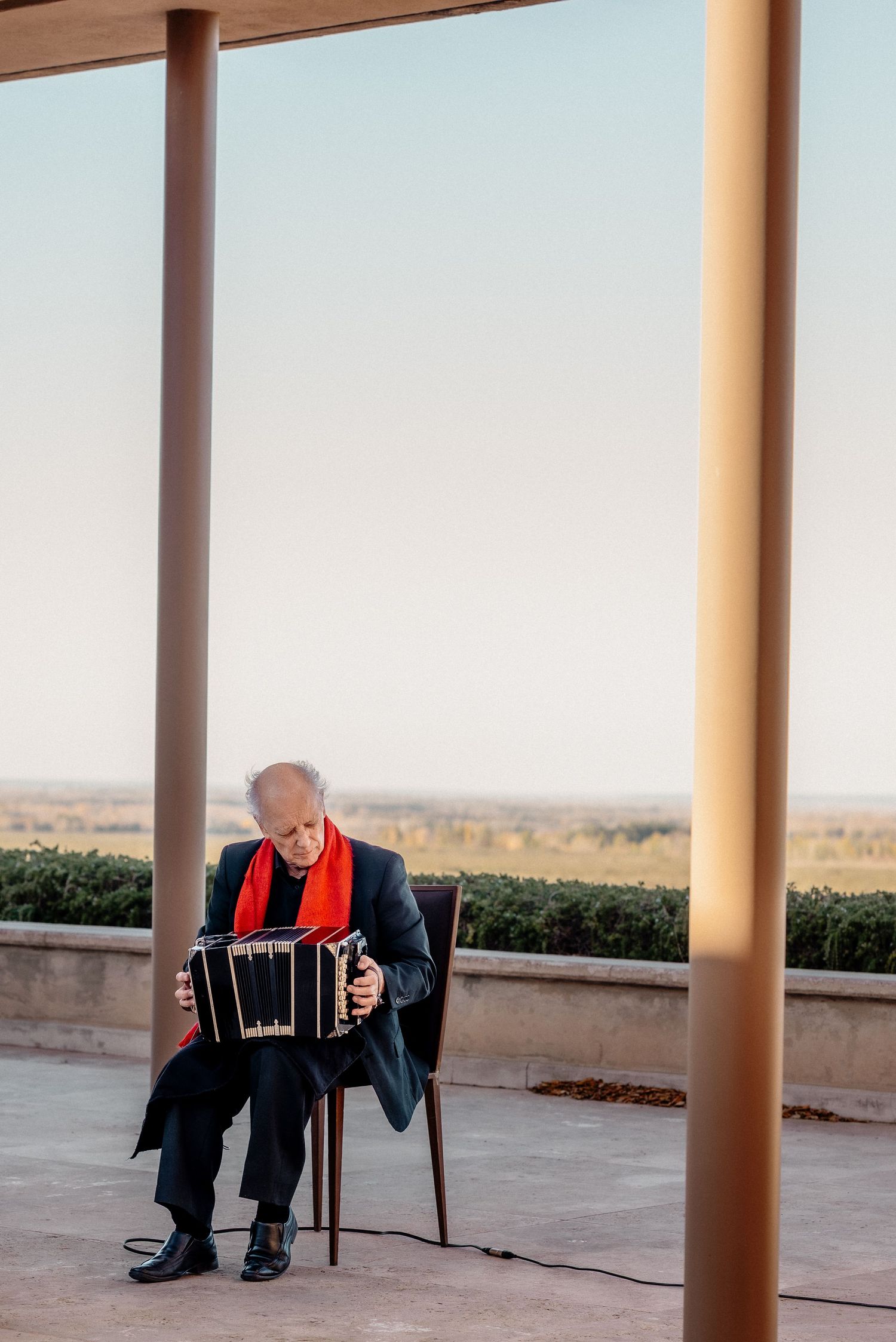 Elderly man playing bandoneon outdoors on terrace, wearing red scarf and dark suit, with vineyard landscape at sunset.