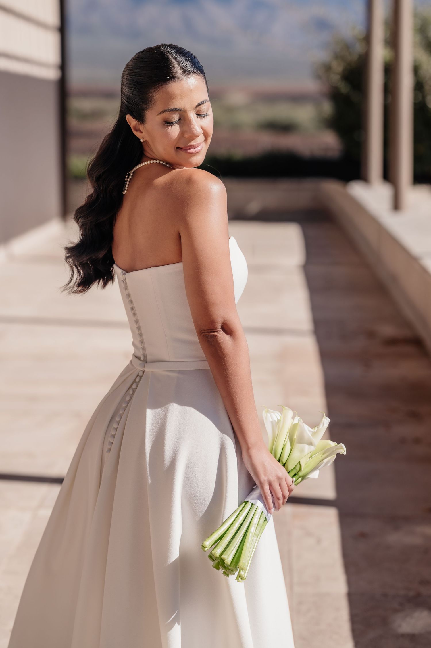 A bride in a strapless white gown holds white calla lilies, smiling softly outdoors on her wedding day.