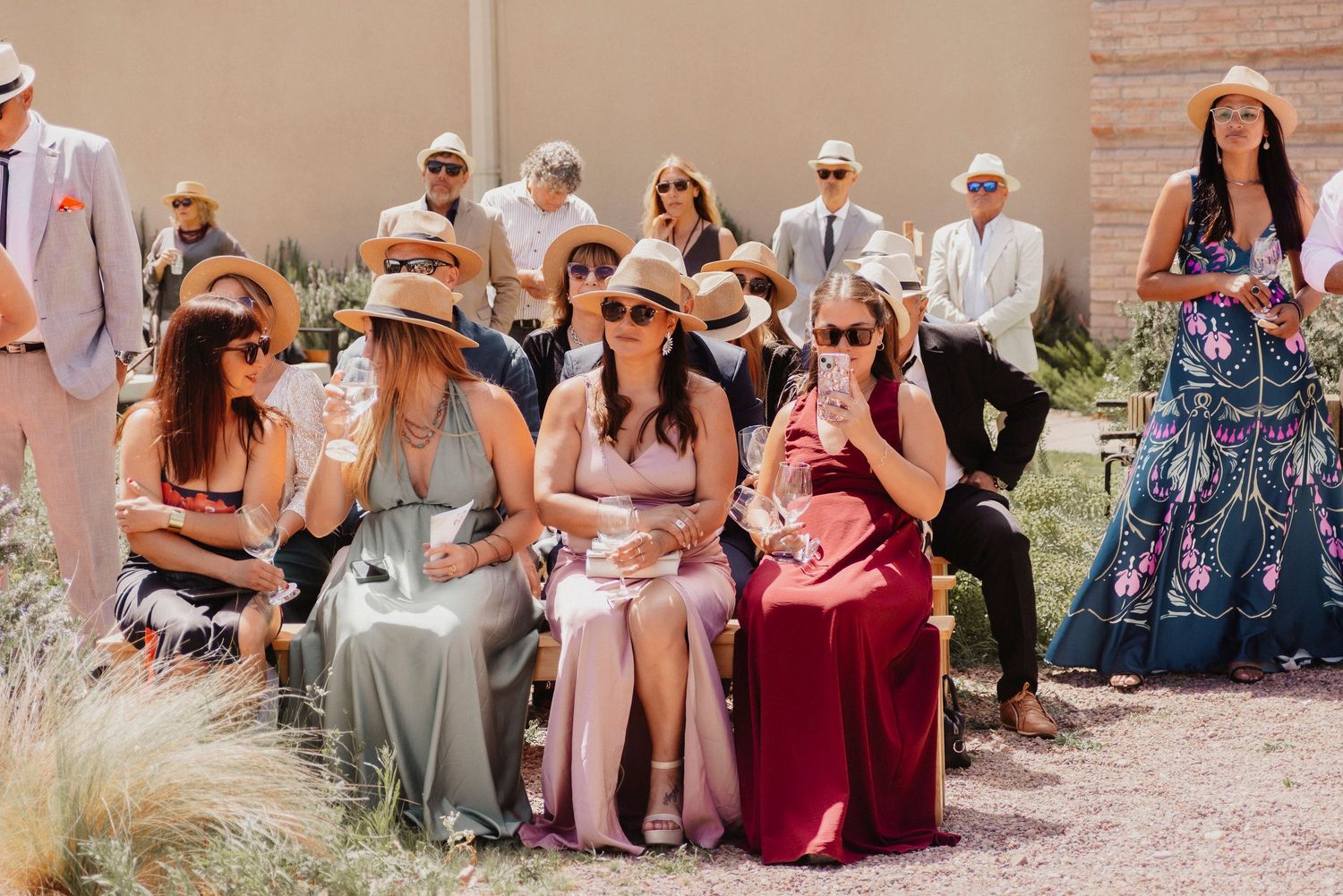 Wedding guests in summer attire sit outdoors watching ceremony with expressions of joy and emotion.