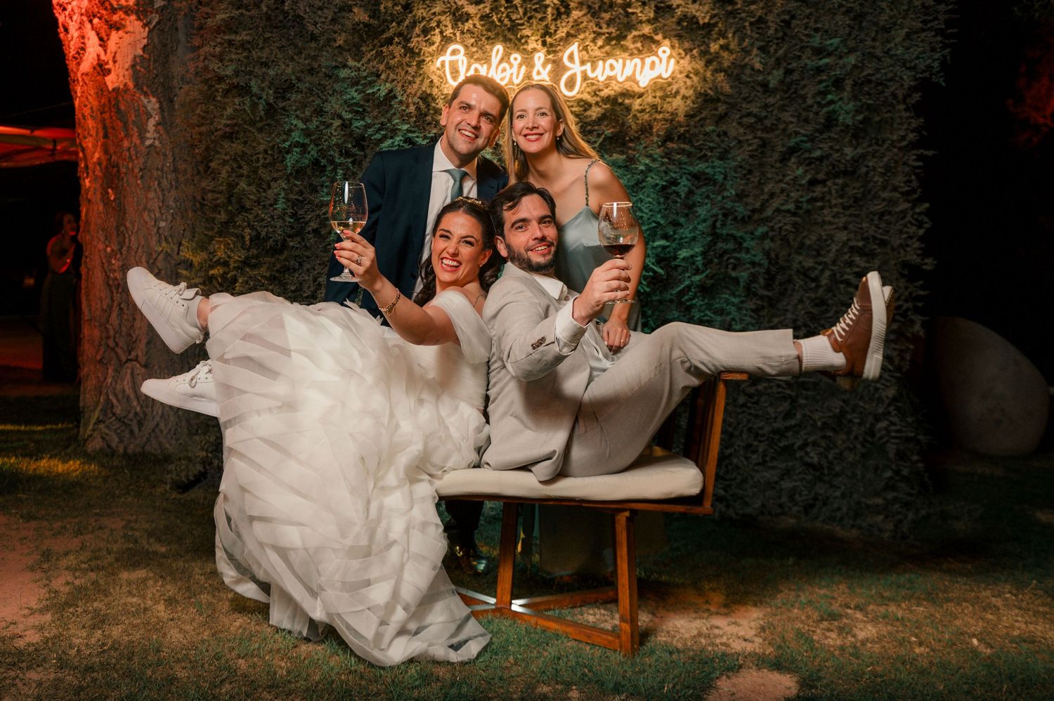 A joyful wedding celebration at night with people posing playfully on a wooden bench under illuminated greenery. fotografos de boda en mendo